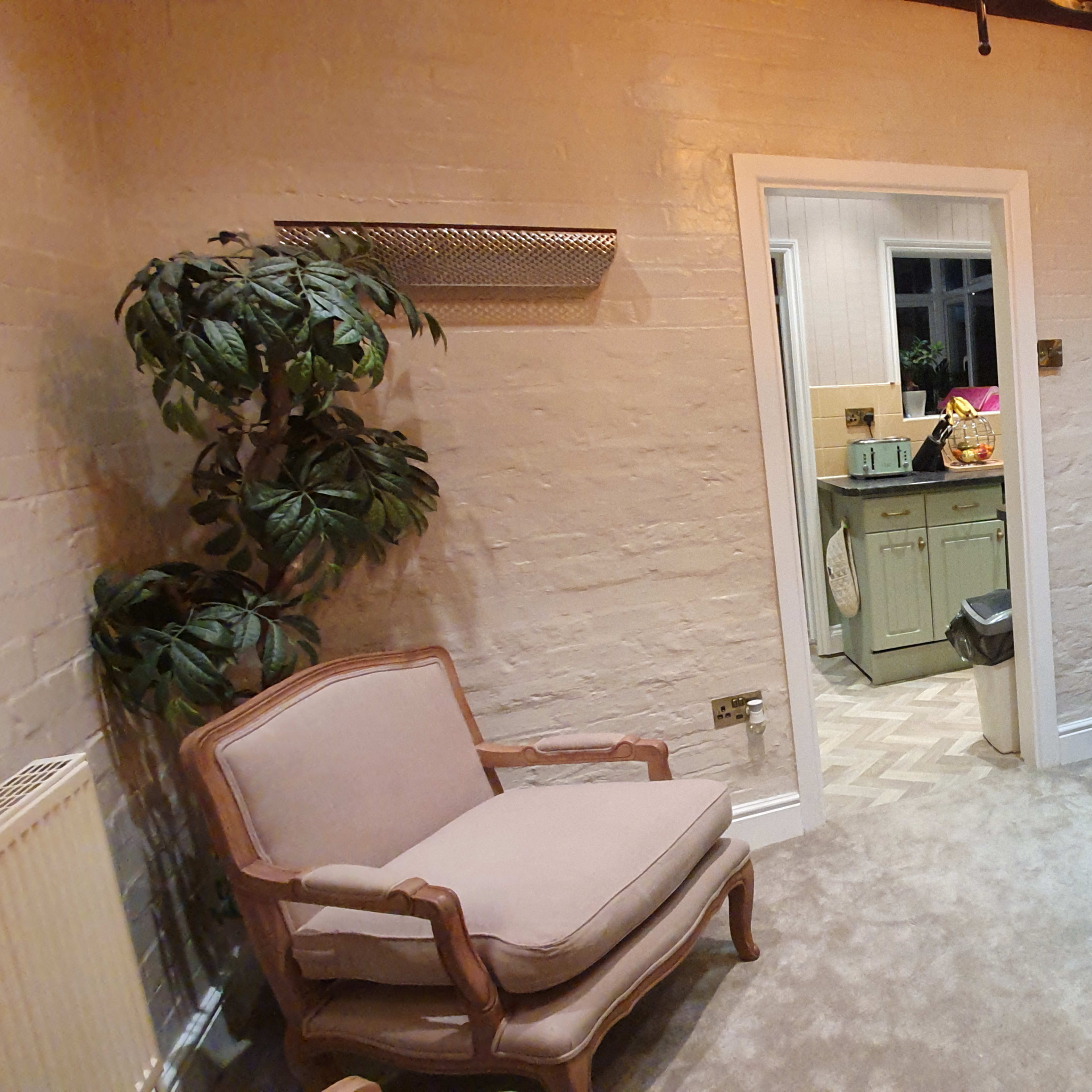 A cozy living room with three beige armchairs arranged around a central area, a fireplace against a brick wall, and a doorway leading to another room.