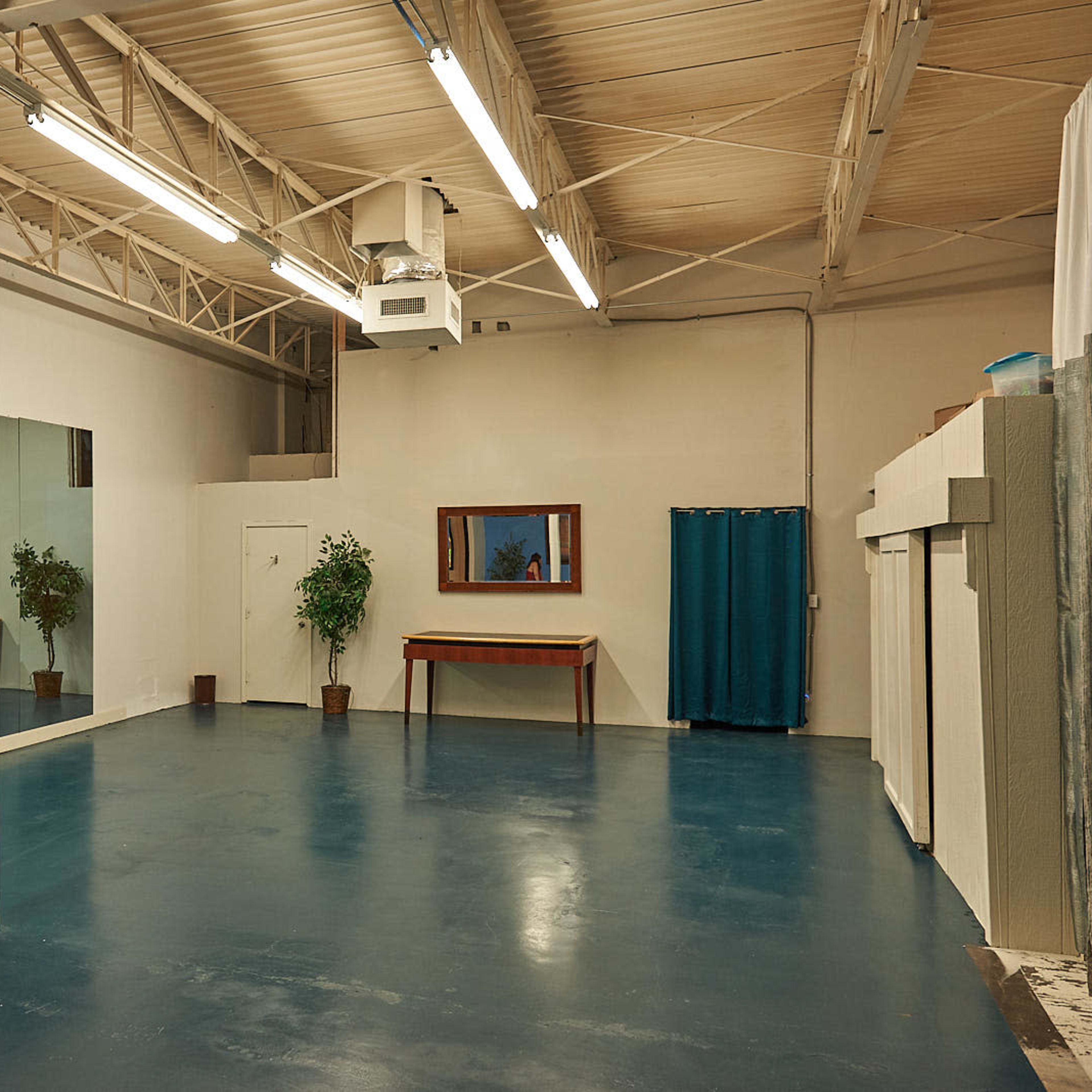 The room features a large mirrored wall, a wooden table against one wall, and a door leading to another space, all set on a glossy blue floor.