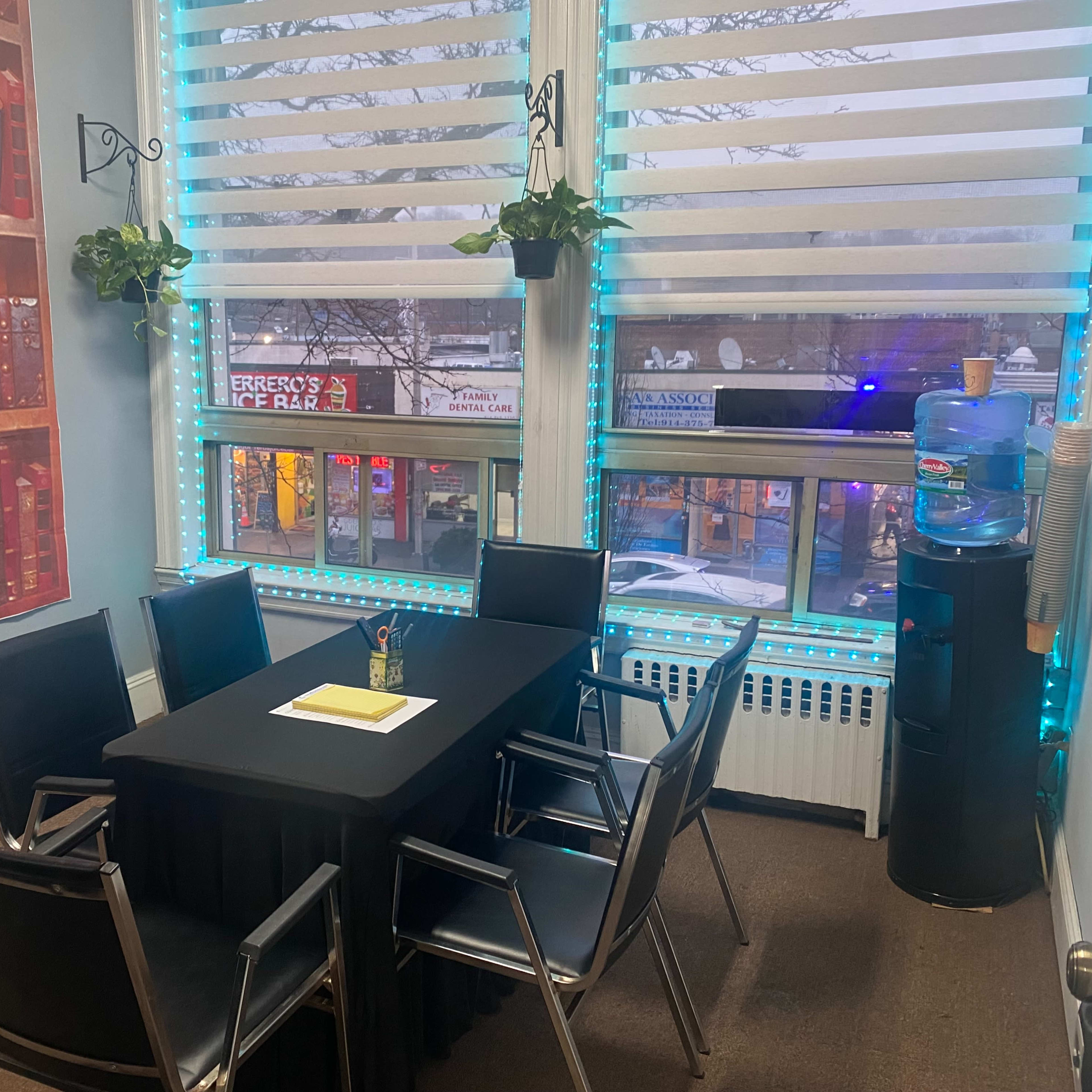 A small meeting room features a black table surrounded by six chairs, with a water cooler and large windows displaying a street view.