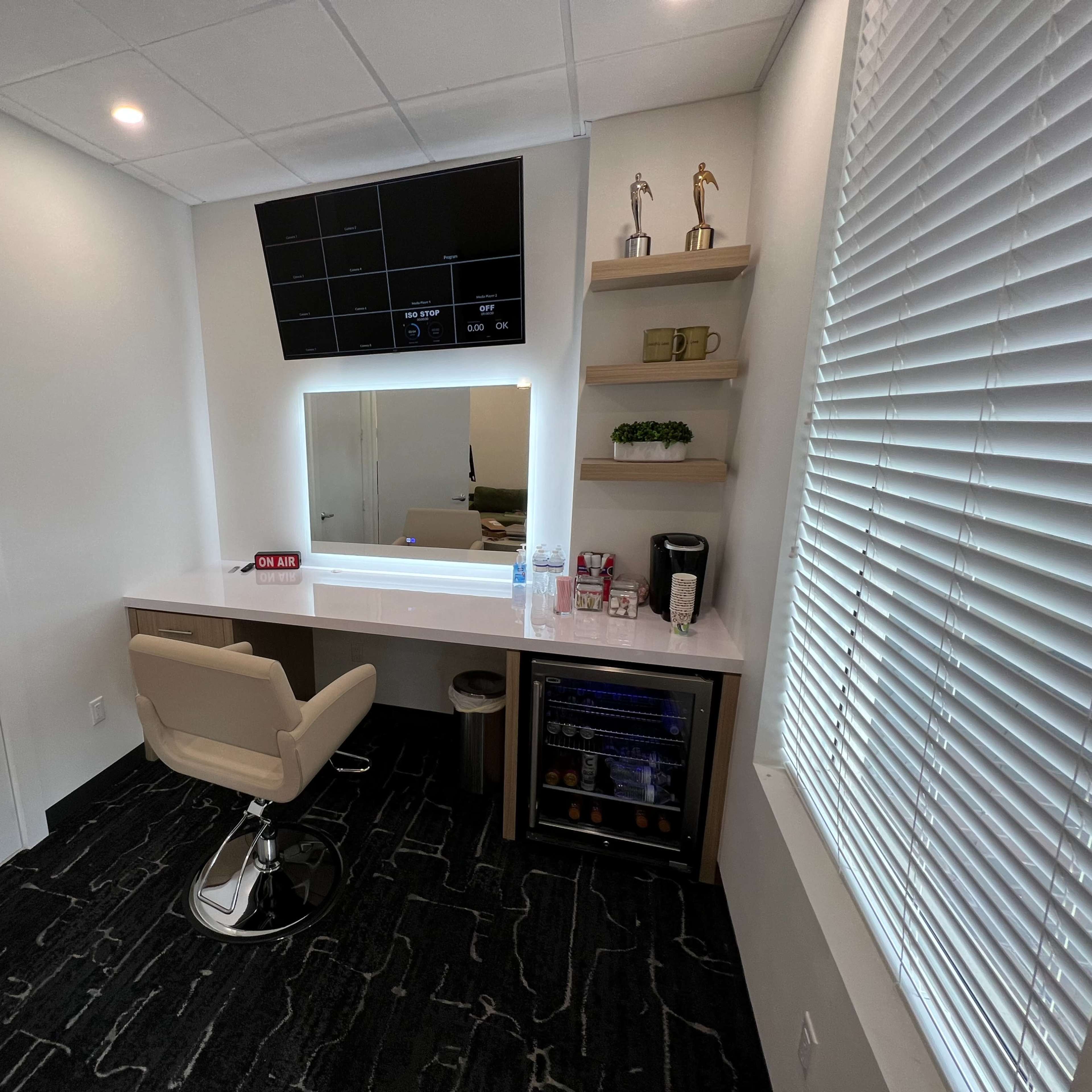 A modern vanity area with a large mirror, a comfortable chair, and a small bar fridge against a wall with shelves displaying items.