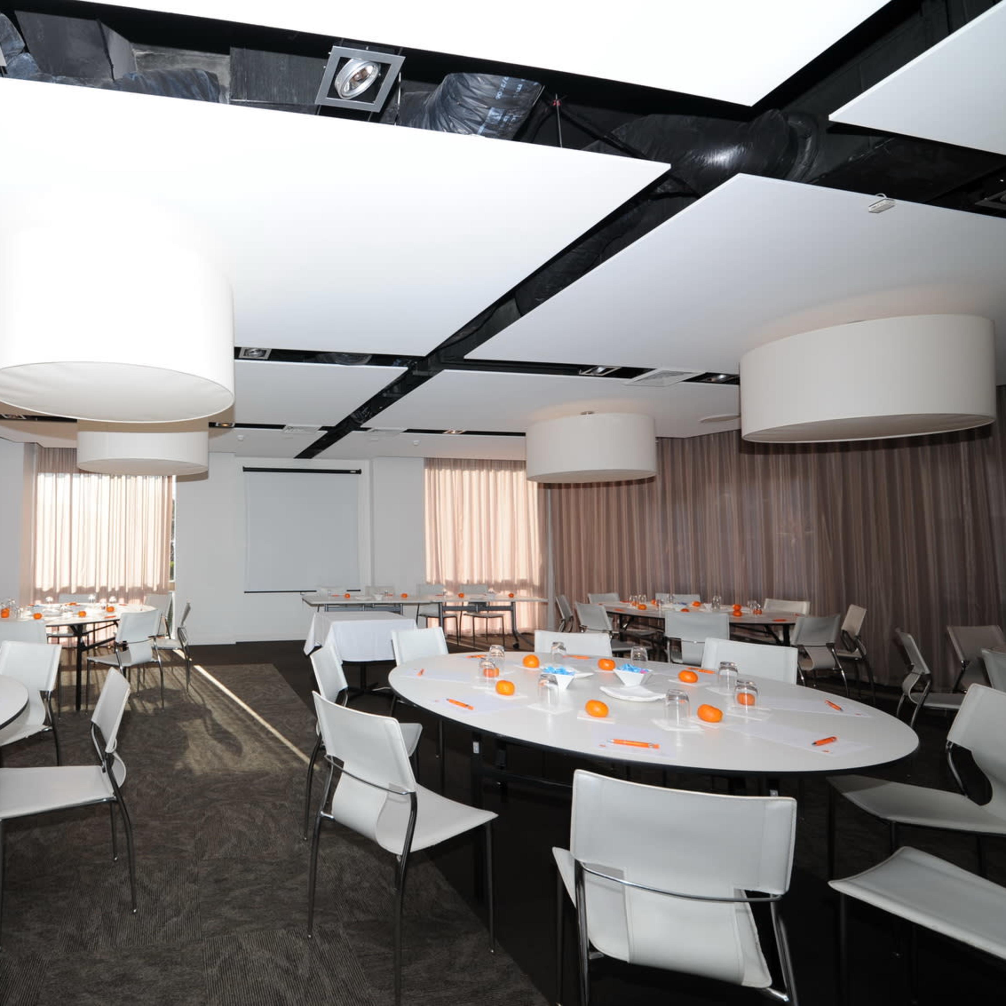 A spacious conference room features several round tables with white tablecloths, orange centerpieces, and large pendant lights hanging from the ceiling.