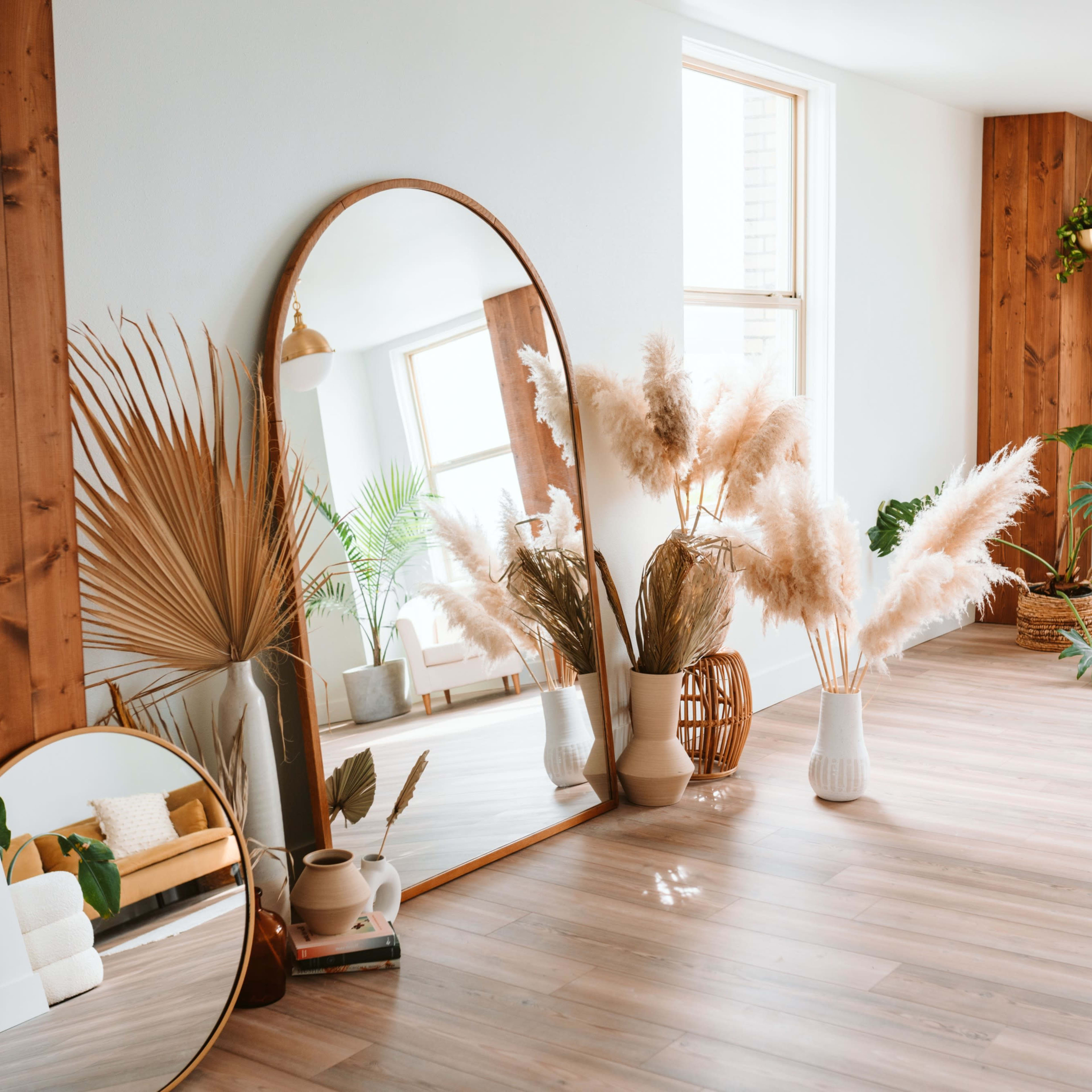 A bright room features large mirrors, decorative plants, and pampas grass arranged near wooden walls and large windows.