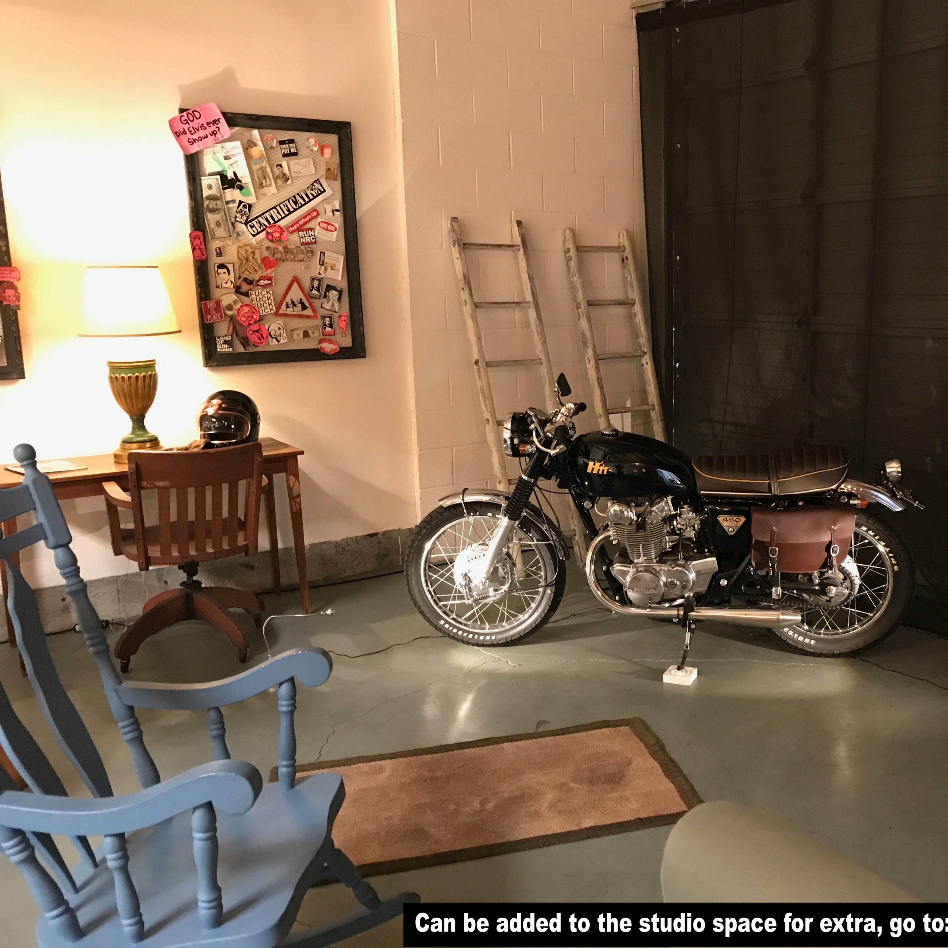 A motorcycle is parked beside a wooden chair and a table in a dimly lit studio space.
