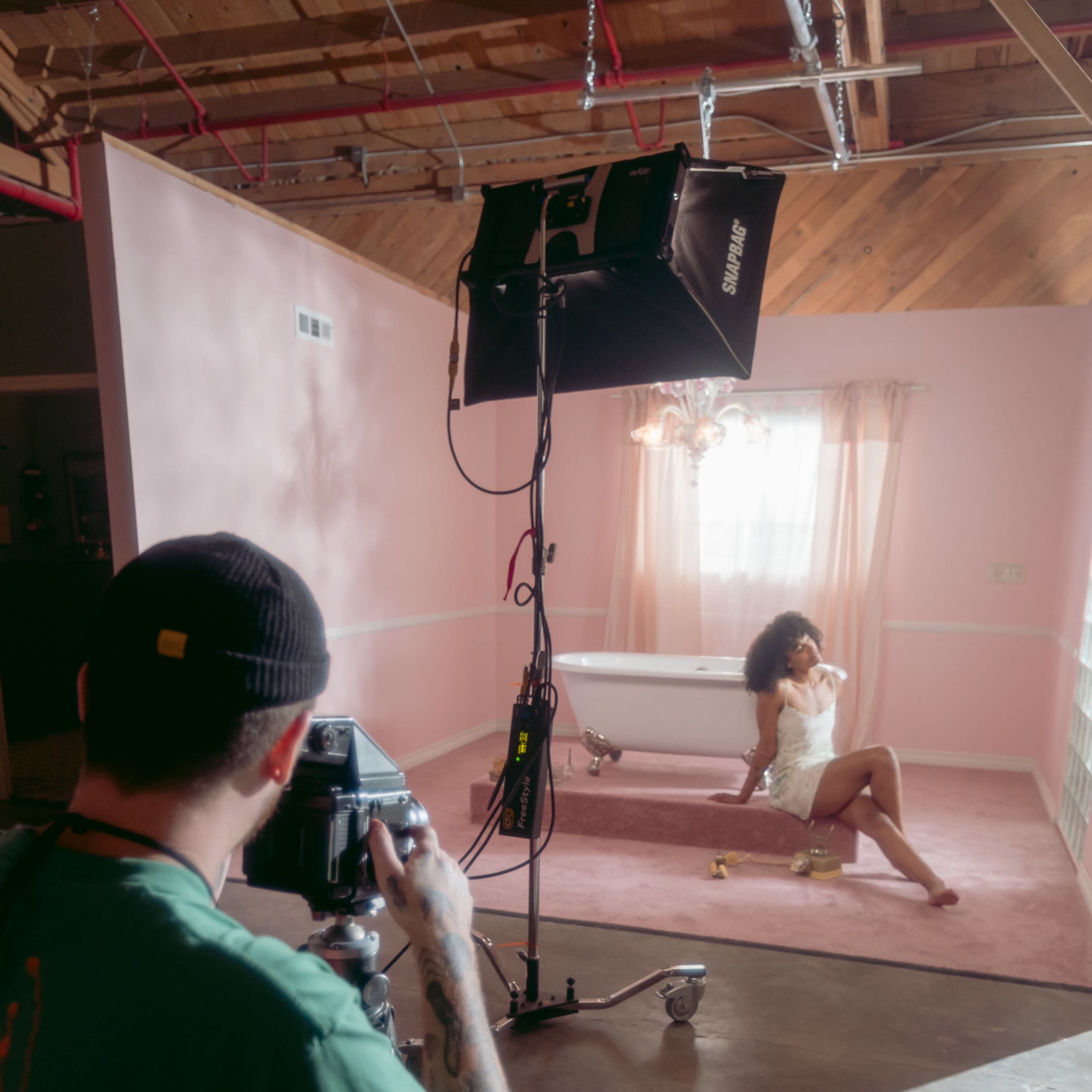 A photographer is taking pictures of a model posed on a rug in a softly lit room with pink walls and a bathtub in the background.