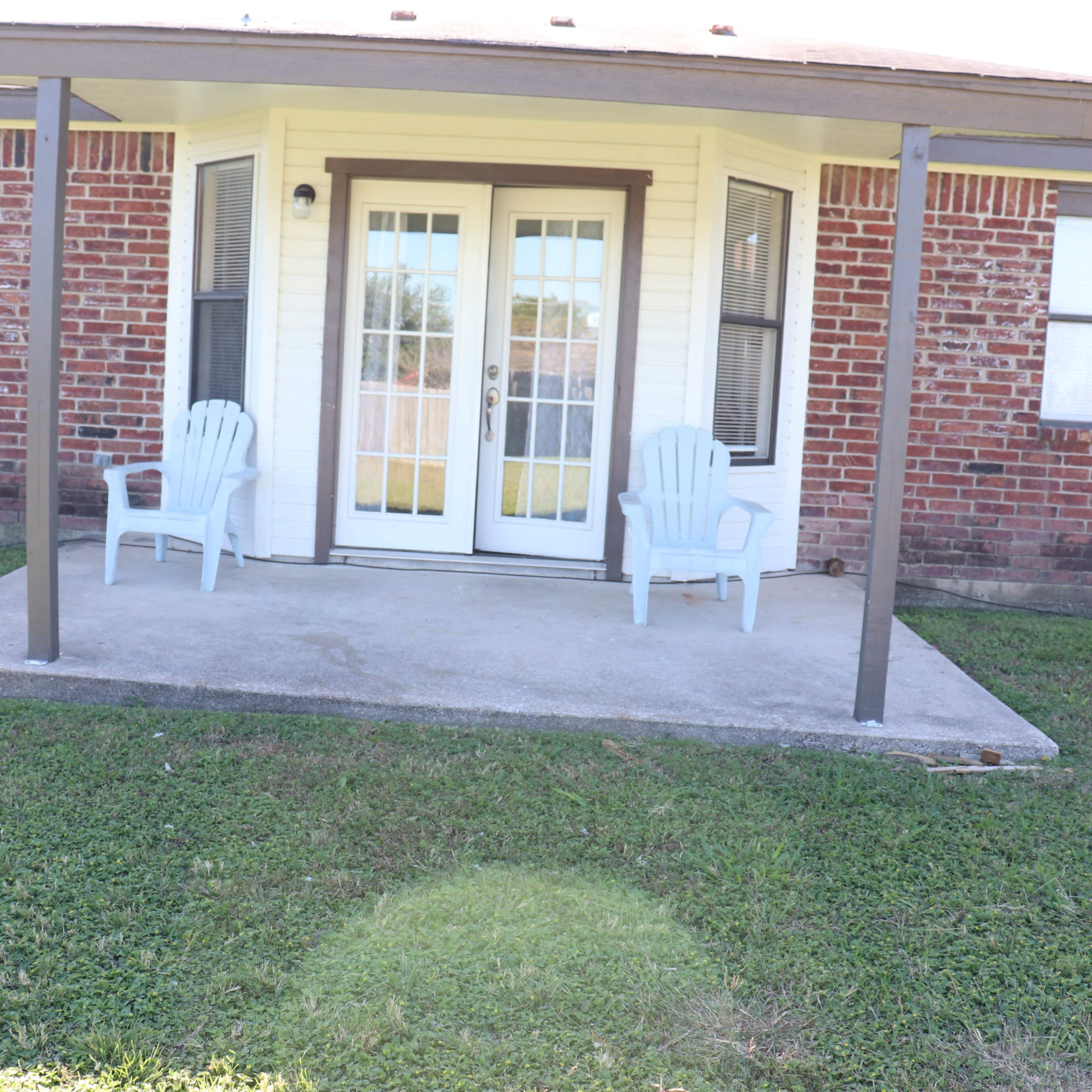 A concrete patio with two white Adirondack chairs in front of a set of double glass doors on a brick house.