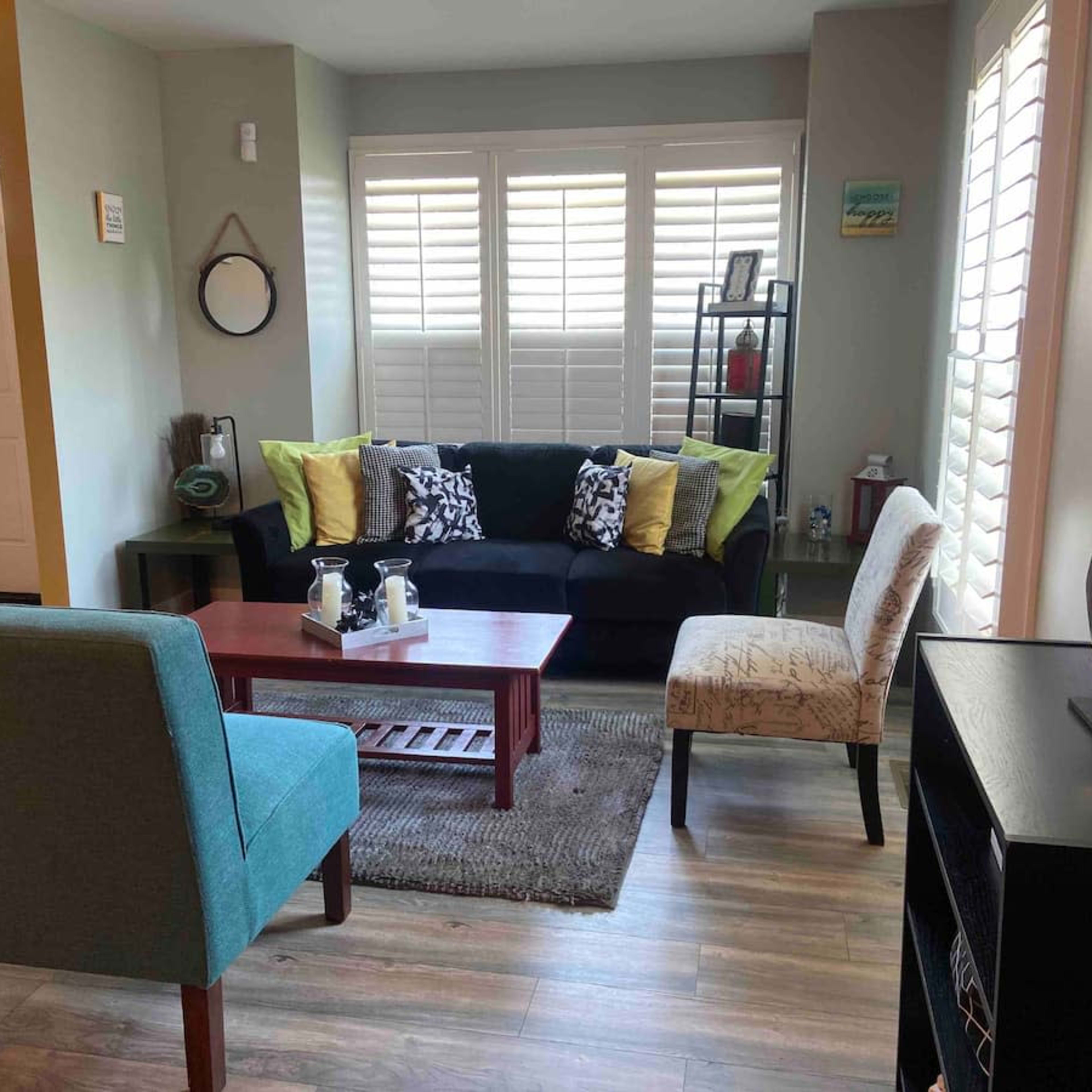 A cozy living room with a black couch, a coffee table, two chairs, and a bookshelf against the wall, all arranged on a wooden floor with window shutters.