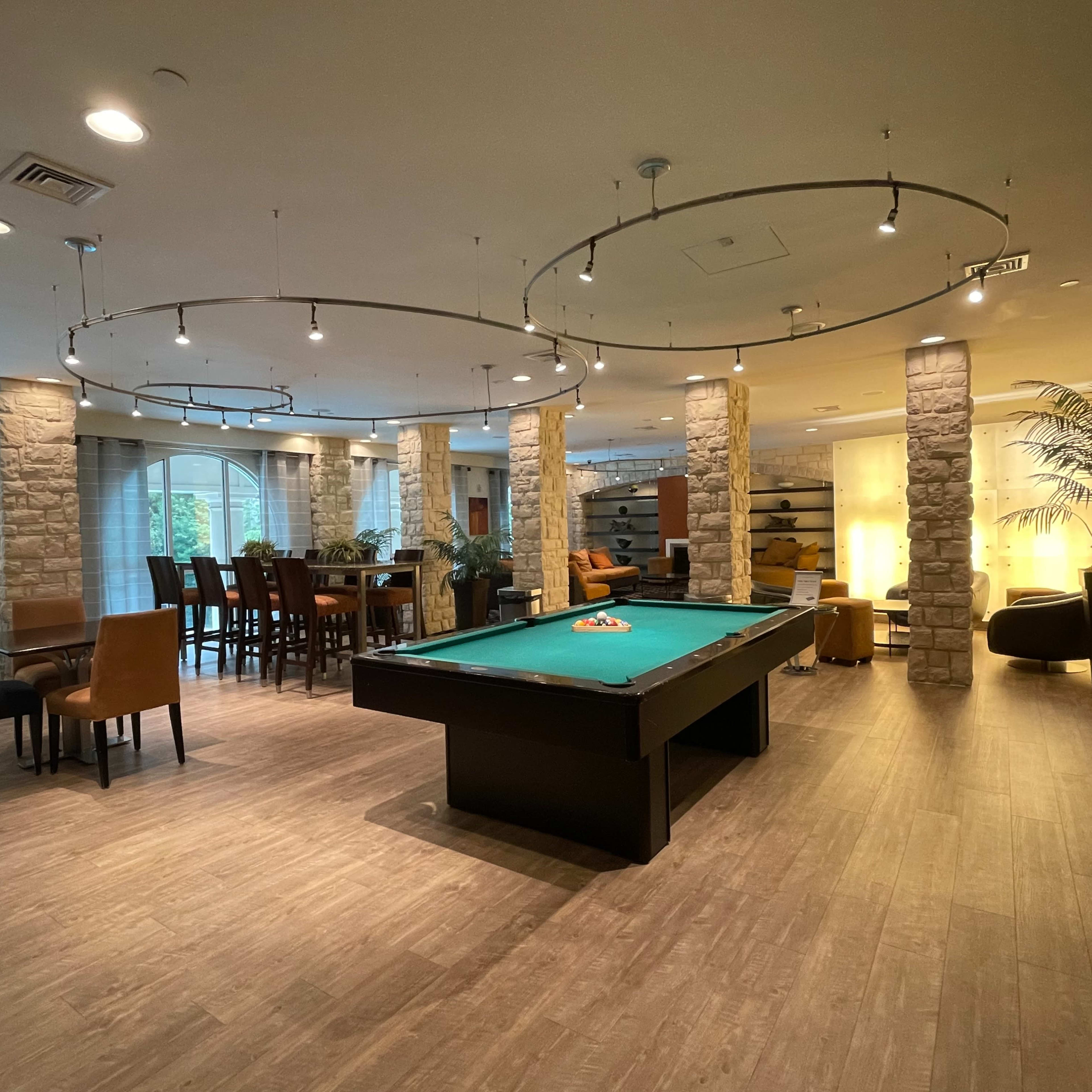 A spacious recreational lounge with a pool table at the center, surrounded by seating areas and illuminated by overhead lights.