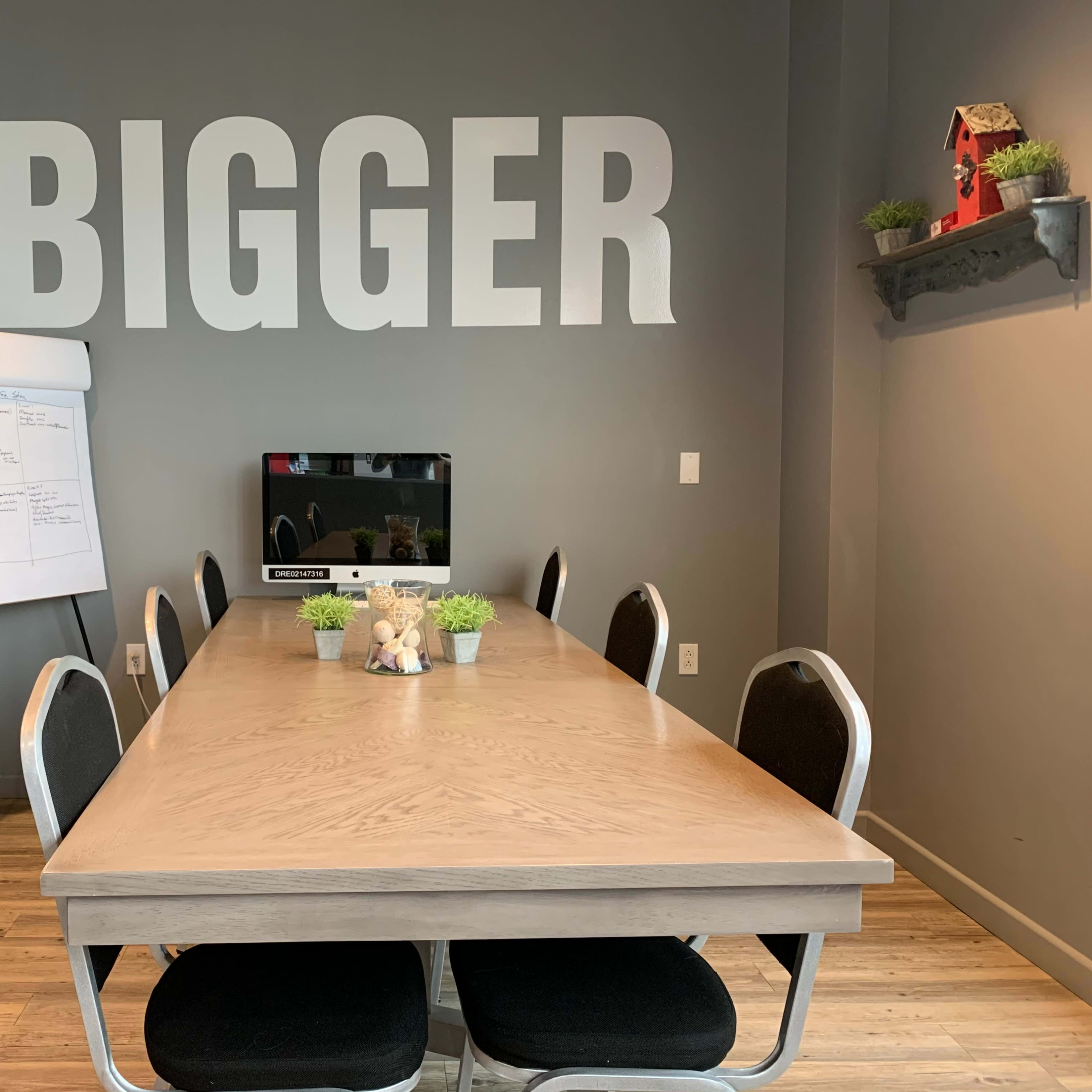 A conference room features a large wooden table surrounded by black chairs, a whiteboard on an easel, a television on the wall, and the phrase "THINK BIGGER" displayed prominently on the back wall.