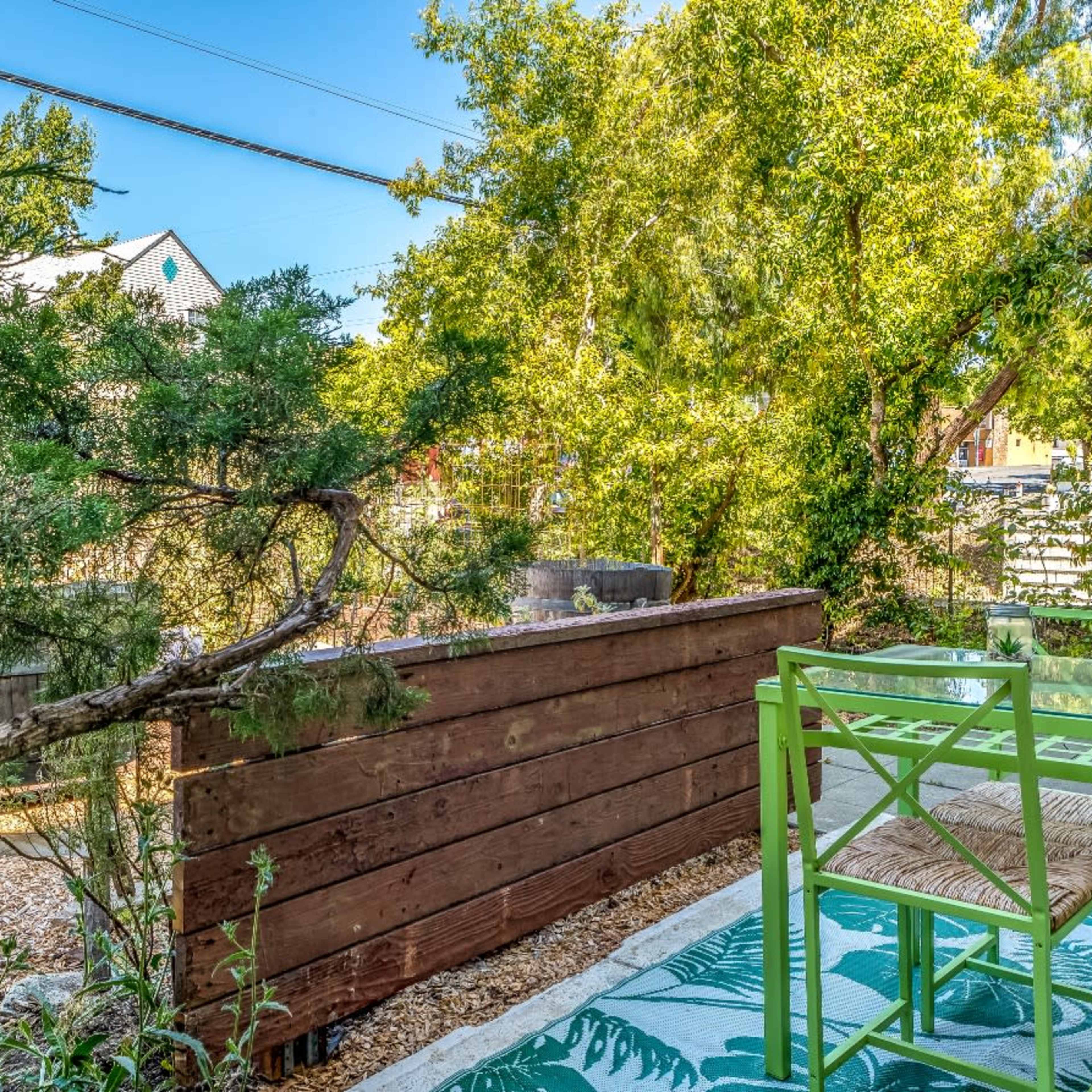 A small outdoor seating area features a green table and chair set surrounded by trees and a wooden fence.