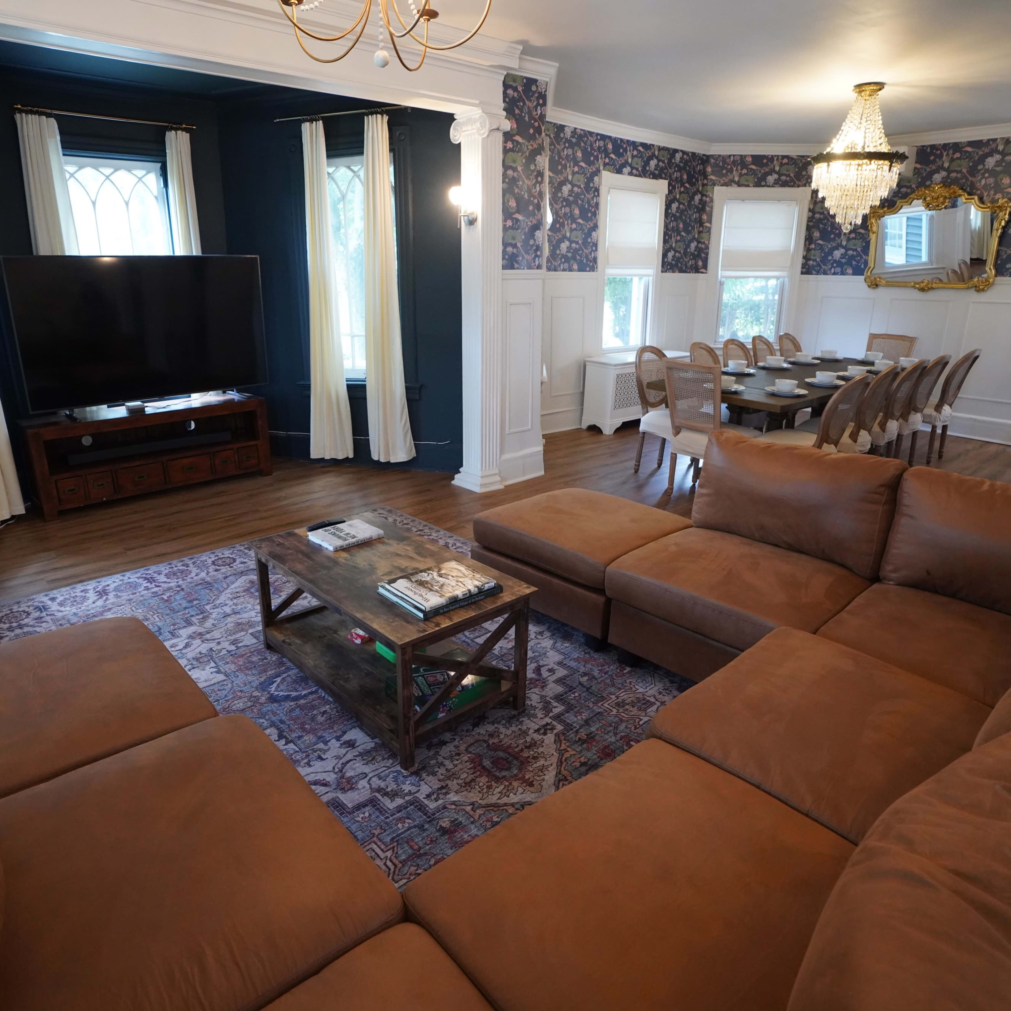 A spacious living room features a brown sectional sofa, a coffee table, a large television, and a dining area with a long table surrounded by chairs.