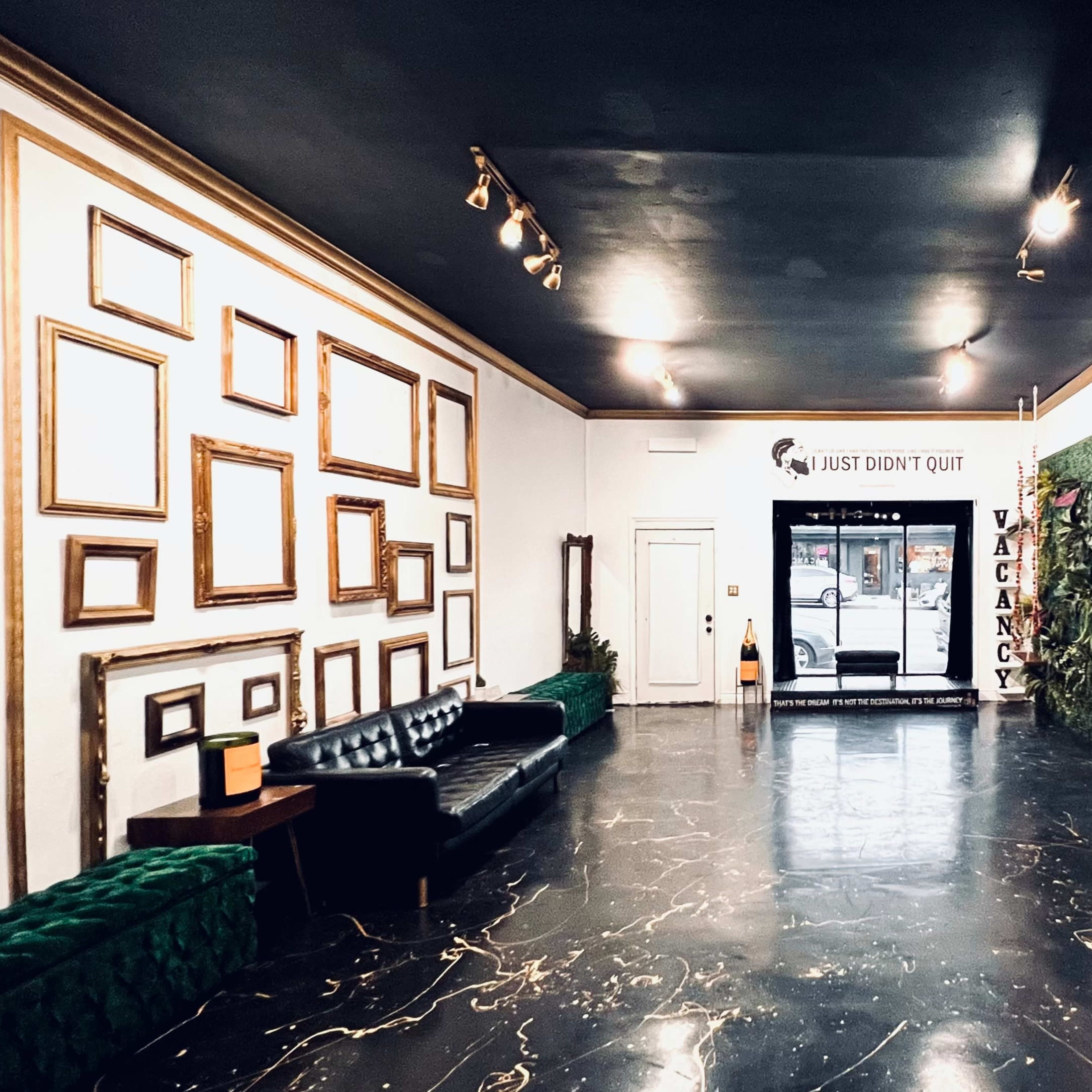 A spacious lobby with a black floor, framed artworks on the walls, a green velvet bench, a black leather sofa, and a doorway leading outside.