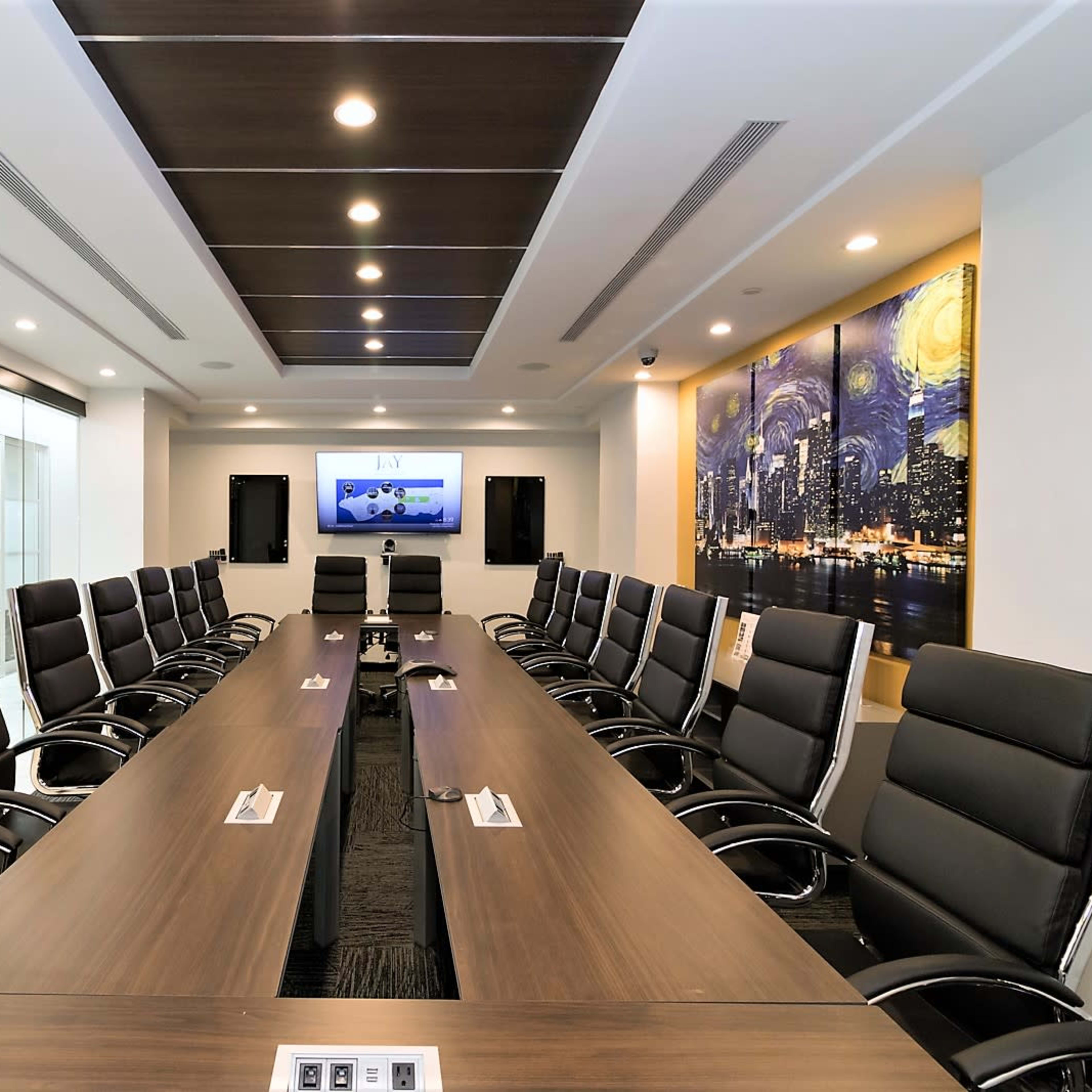 A modern conference room features a long wooden table surrounded by black leather chairs, with a large screen and a cityscape painting on the wall.
