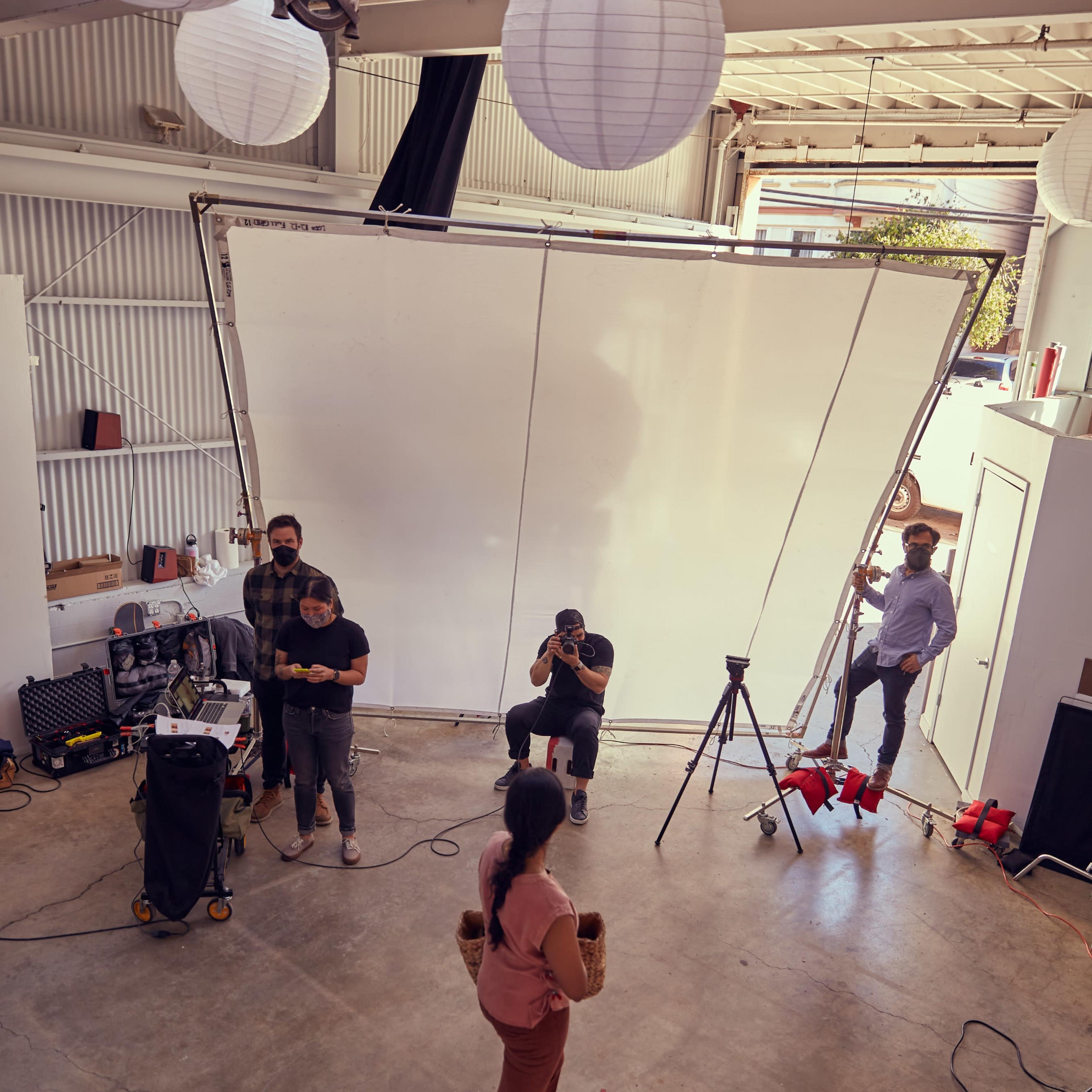 A photoshoot is taking place in a spacious studio with a model posing in front of a large backdrop while photographers and crew members are present.