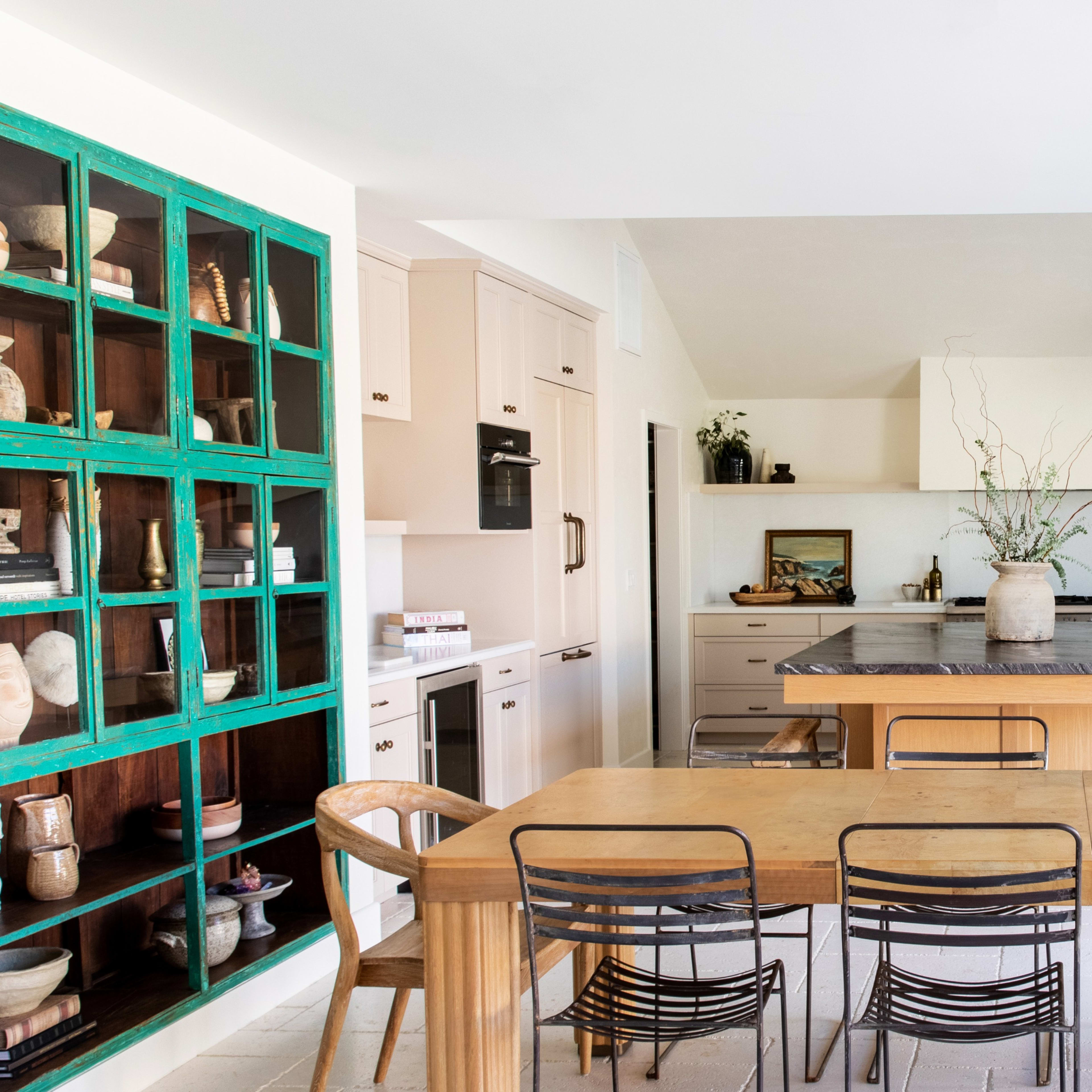 A modern kitchen features a large central island, a wooden dining table with black chairs, and an open shelving unit painted teal displaying ceramic pieces.