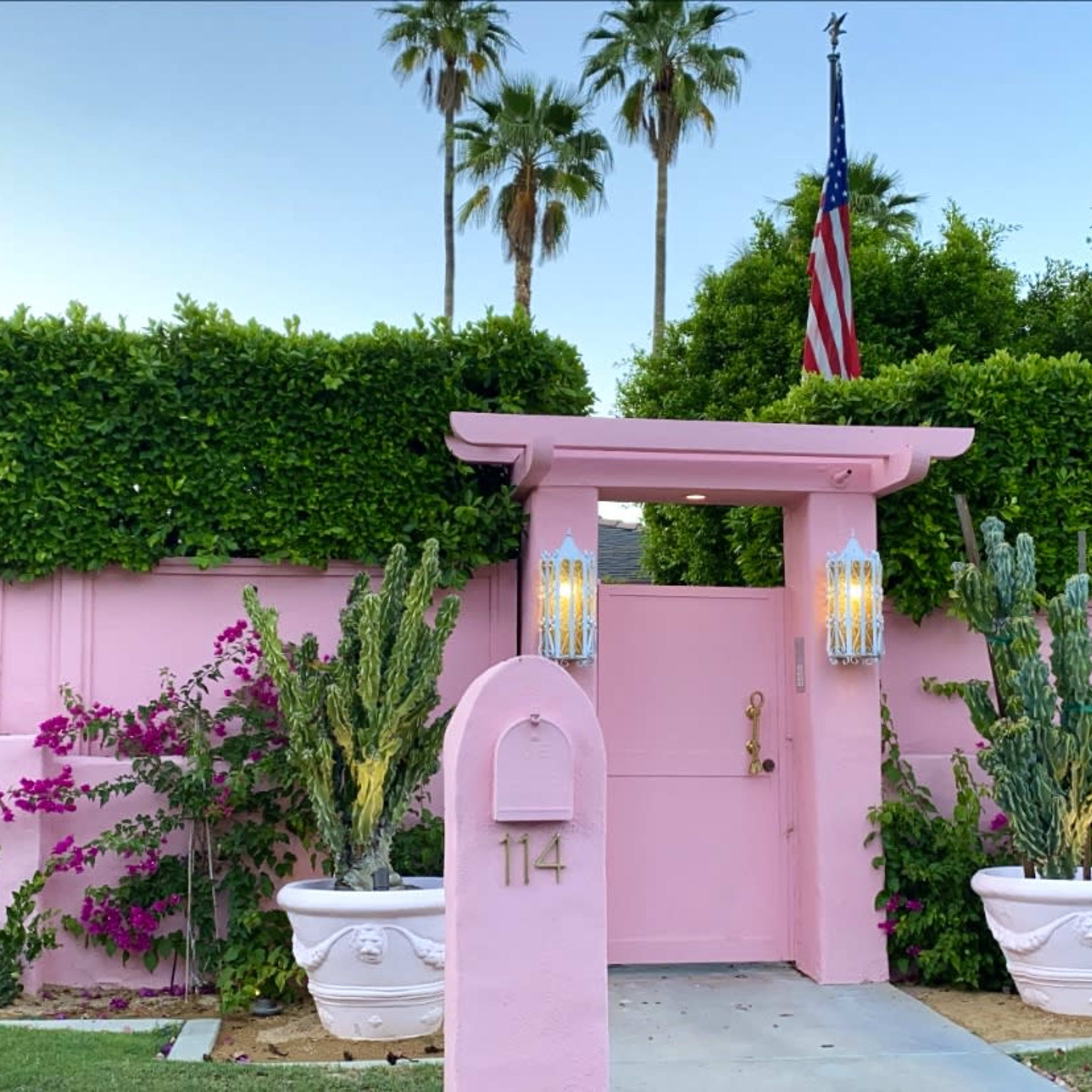 A pink entrance gate adorned with lanterns and surrounded by lush greenery and flowering plants features the number 114 and an American flag in the background.