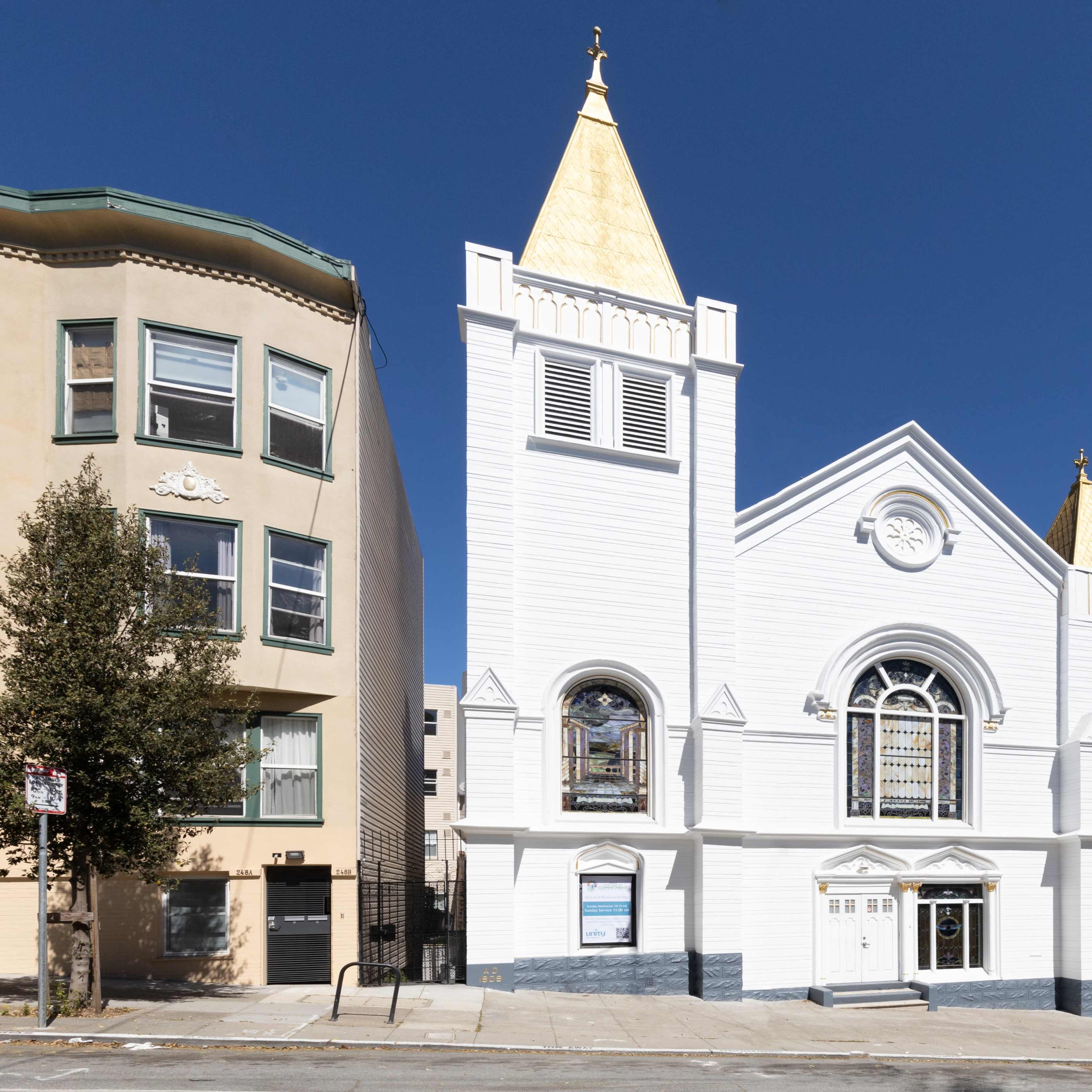A white church with two golden spires situated at the corner of a street, flanked by two residential buildings.