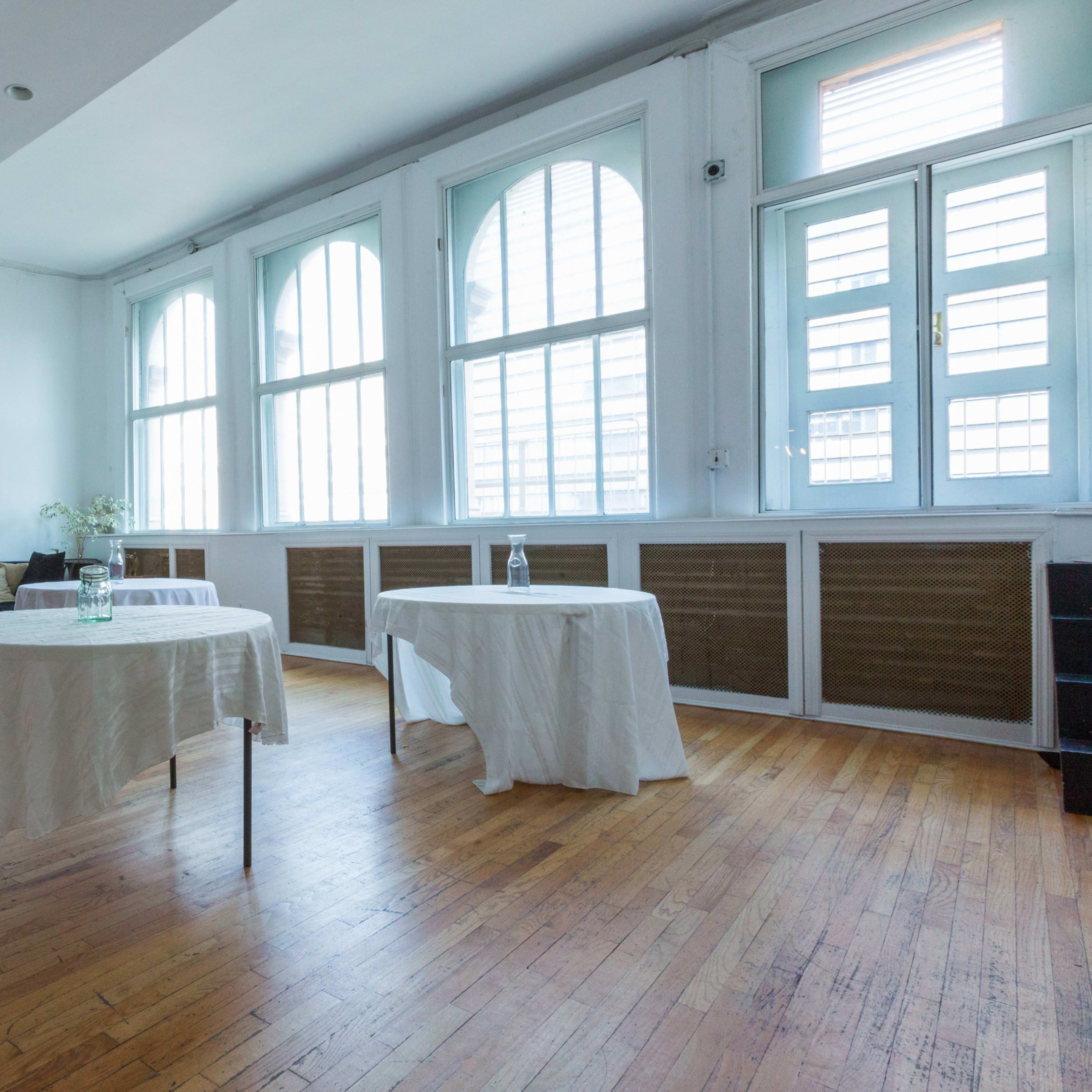 A minimalist room with two small tables covered in white cloths, large windows letting in natural light, and an elevator door on the left.
