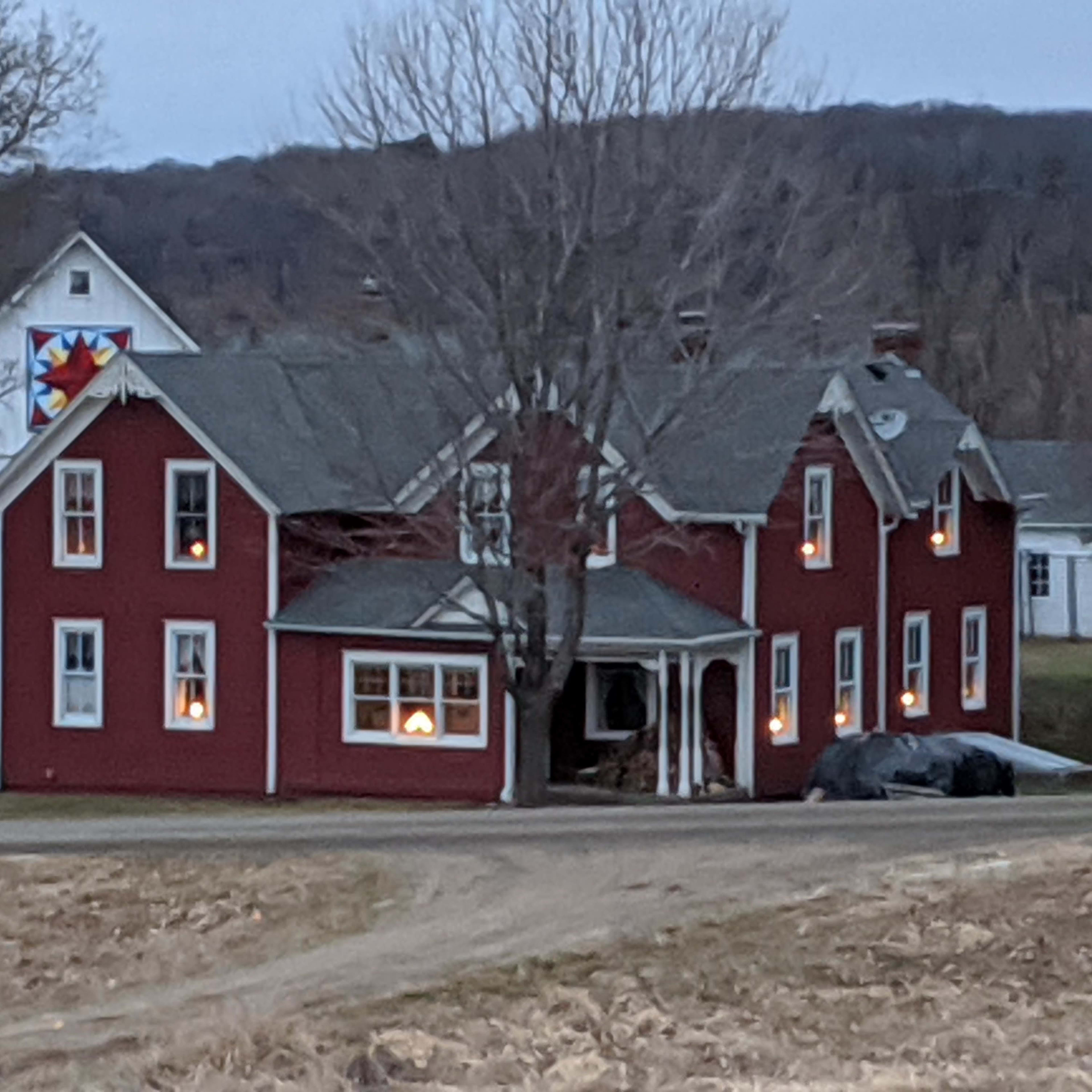 A red farmhouse with illuminated windows and a quilt pattern on the adjoining white building is set against a backdrop of bare trees and a twilight sky.