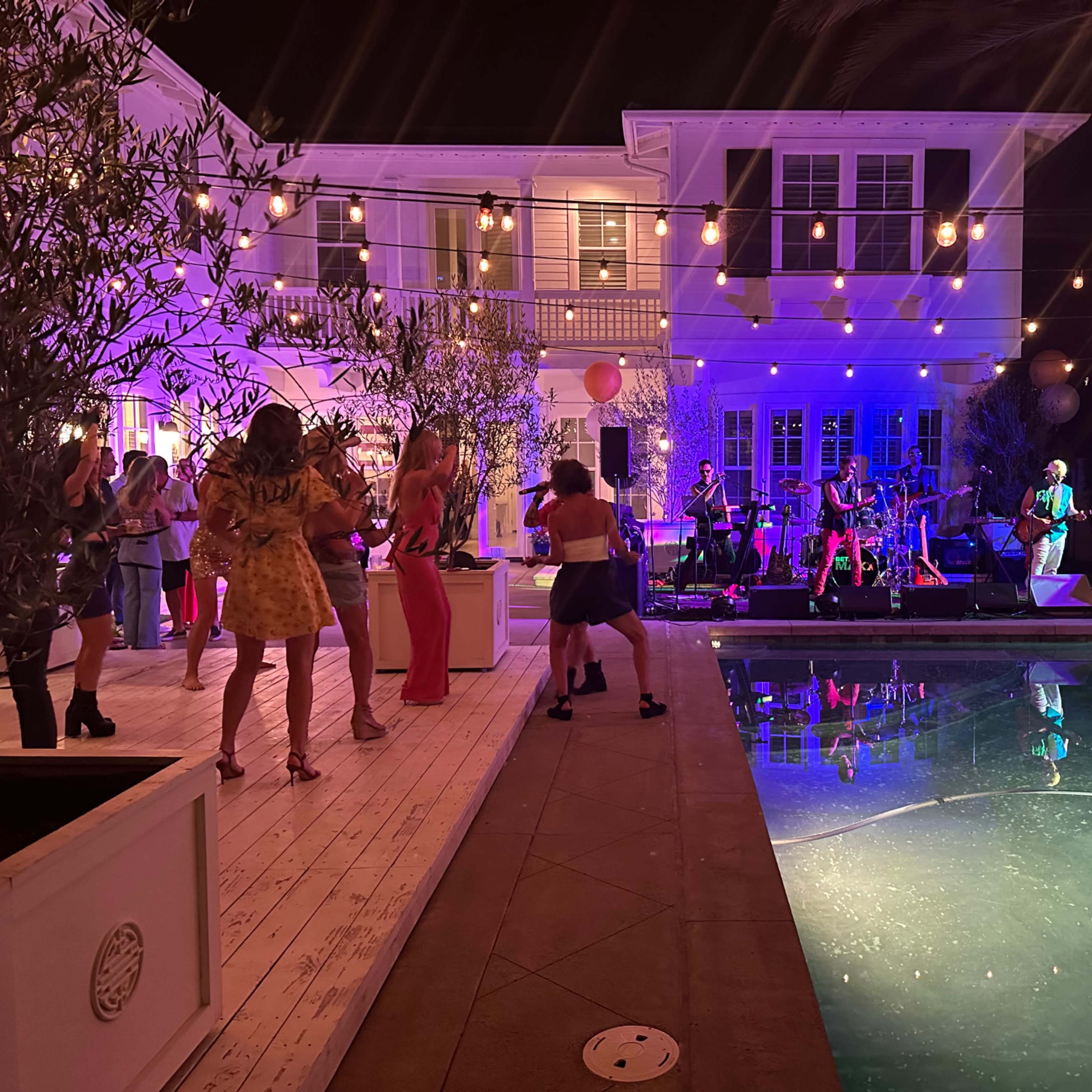 The scene shows a lively outdoor party by a pool at night, with guests dancing and a live band performing under string lights and colorful decorations.