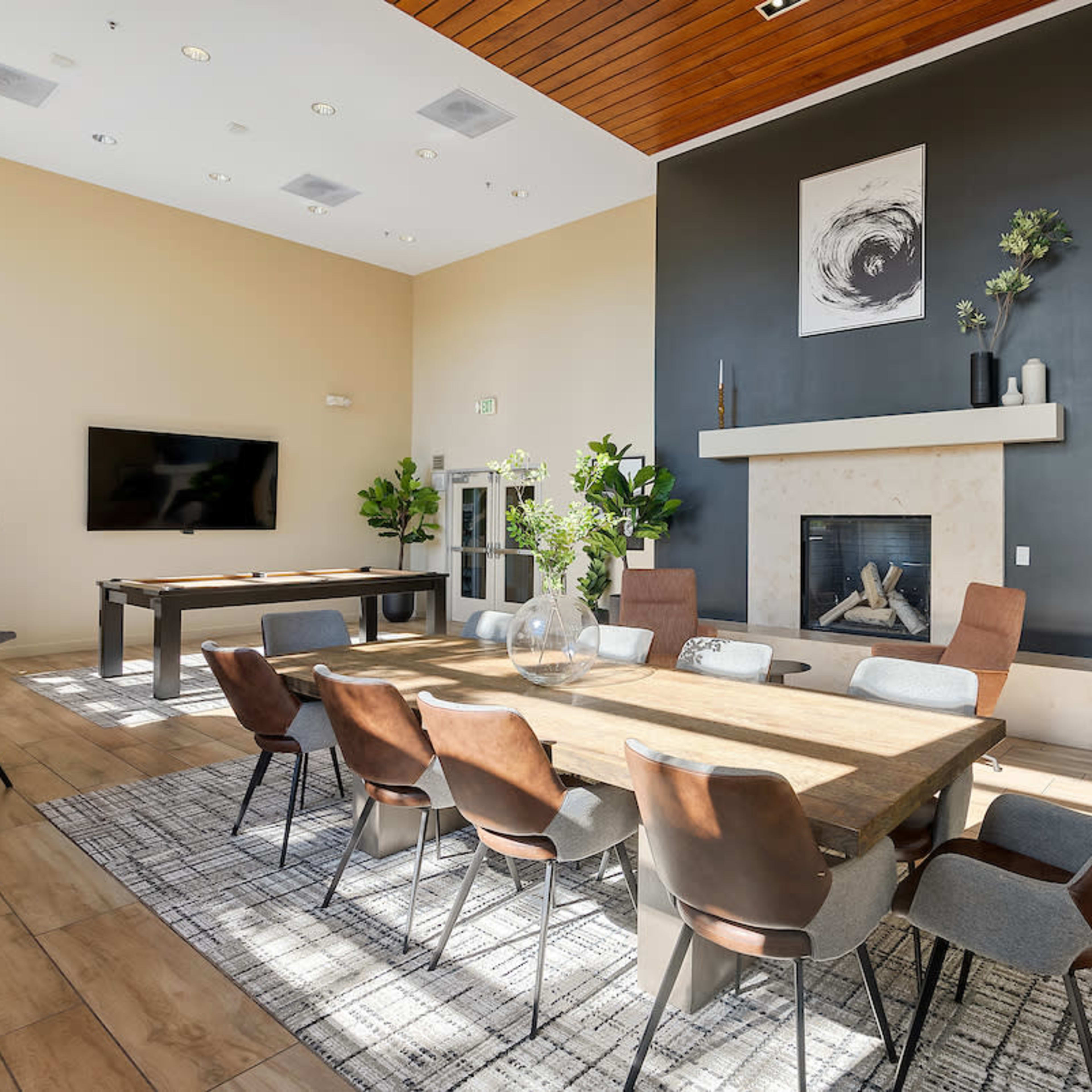 A modern communal room with a large wooden dining table surrounded by various chairs, a fireplace, a television, and potted plants.