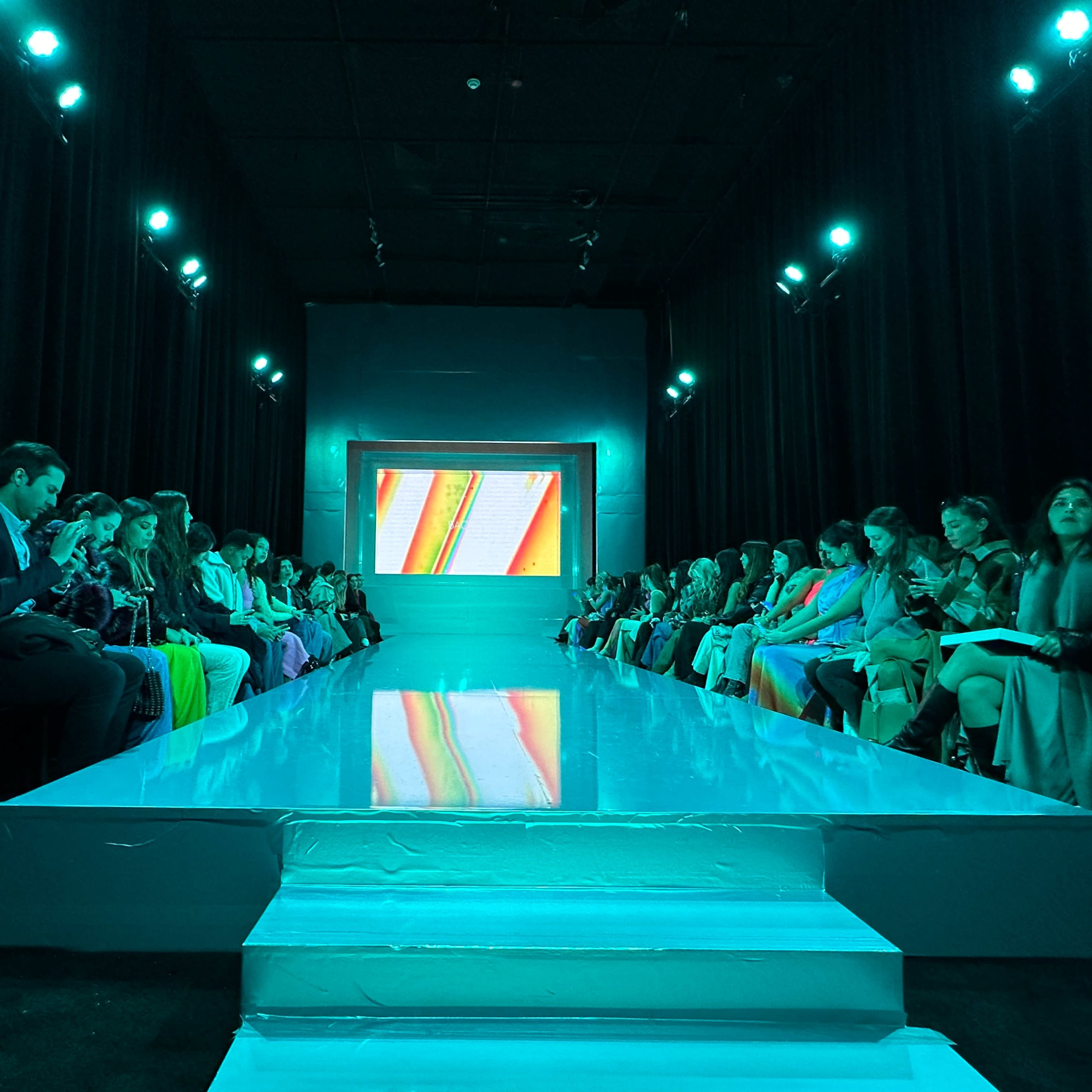 A fashion show runway with a glossy white surface, flanked by rows of seated audience members under teal lighting and a bright screen at the end of the runway.