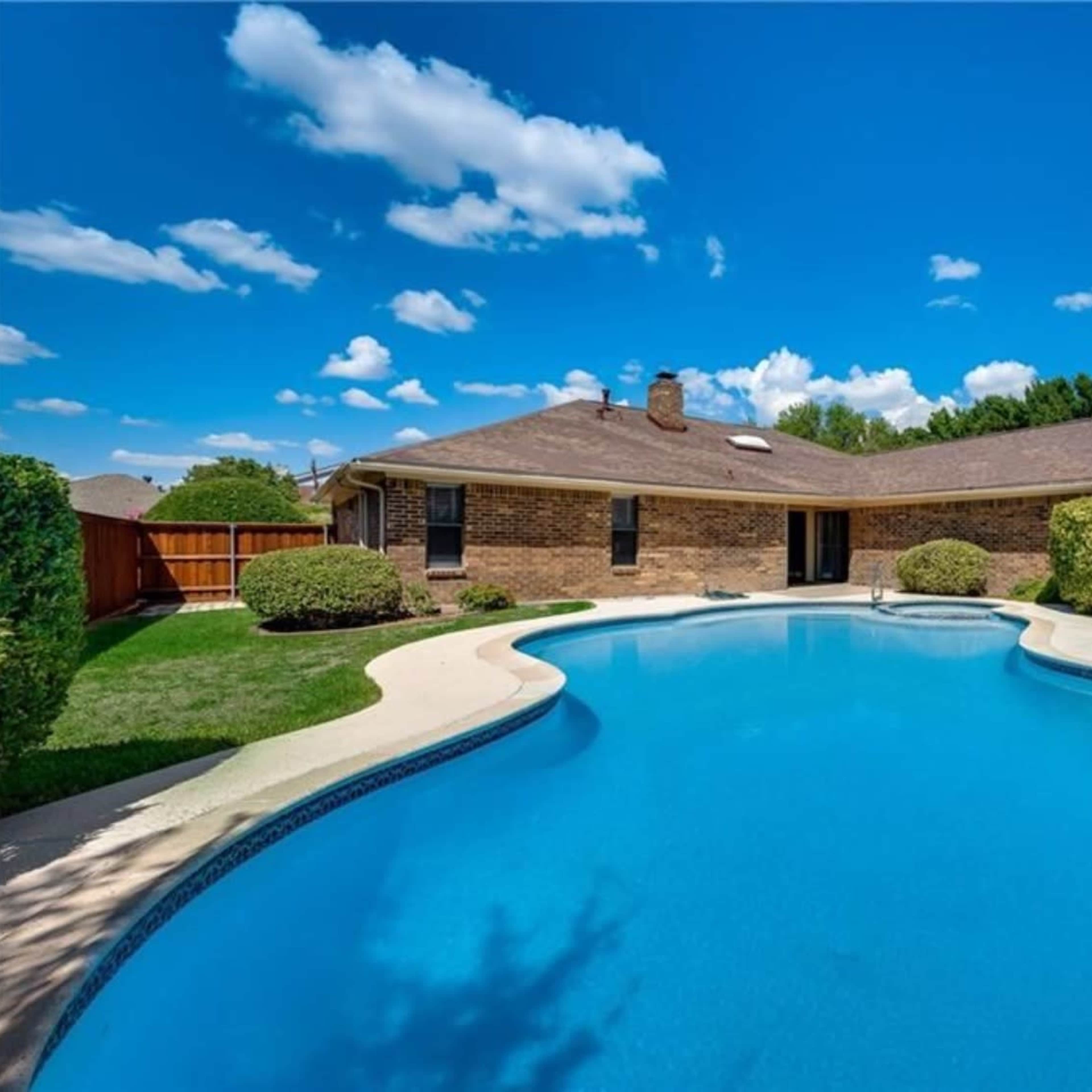 A clear blue swimming pool is surrounded by a well-manicured lawn and bushes, with a single-story brick house in the background under a bright sky with clouds.