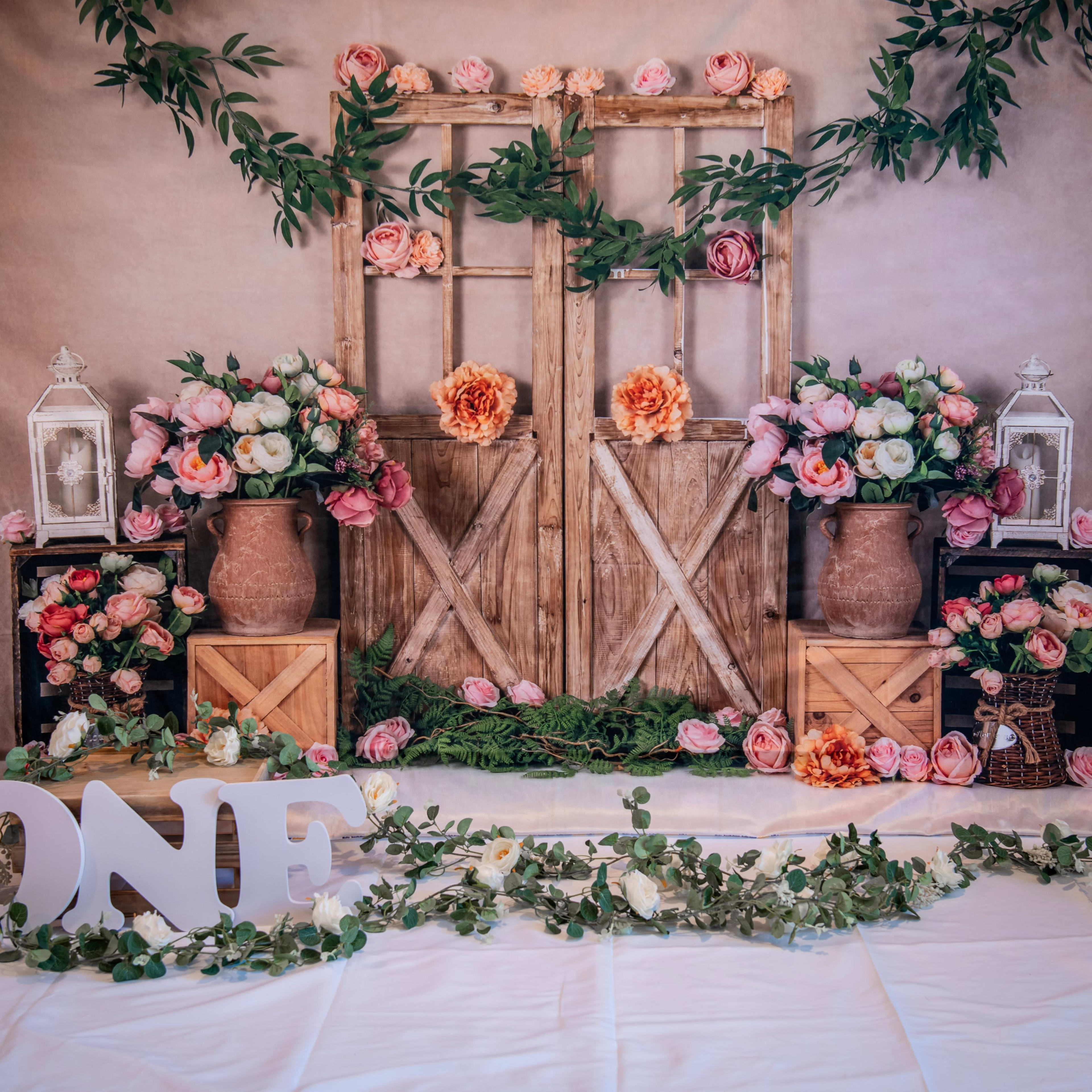 A decorative setup featuring a wooden double door adorned with flowers, surrounded by potted plants and lanterns, with the word "ONE" displayed prominently in front.