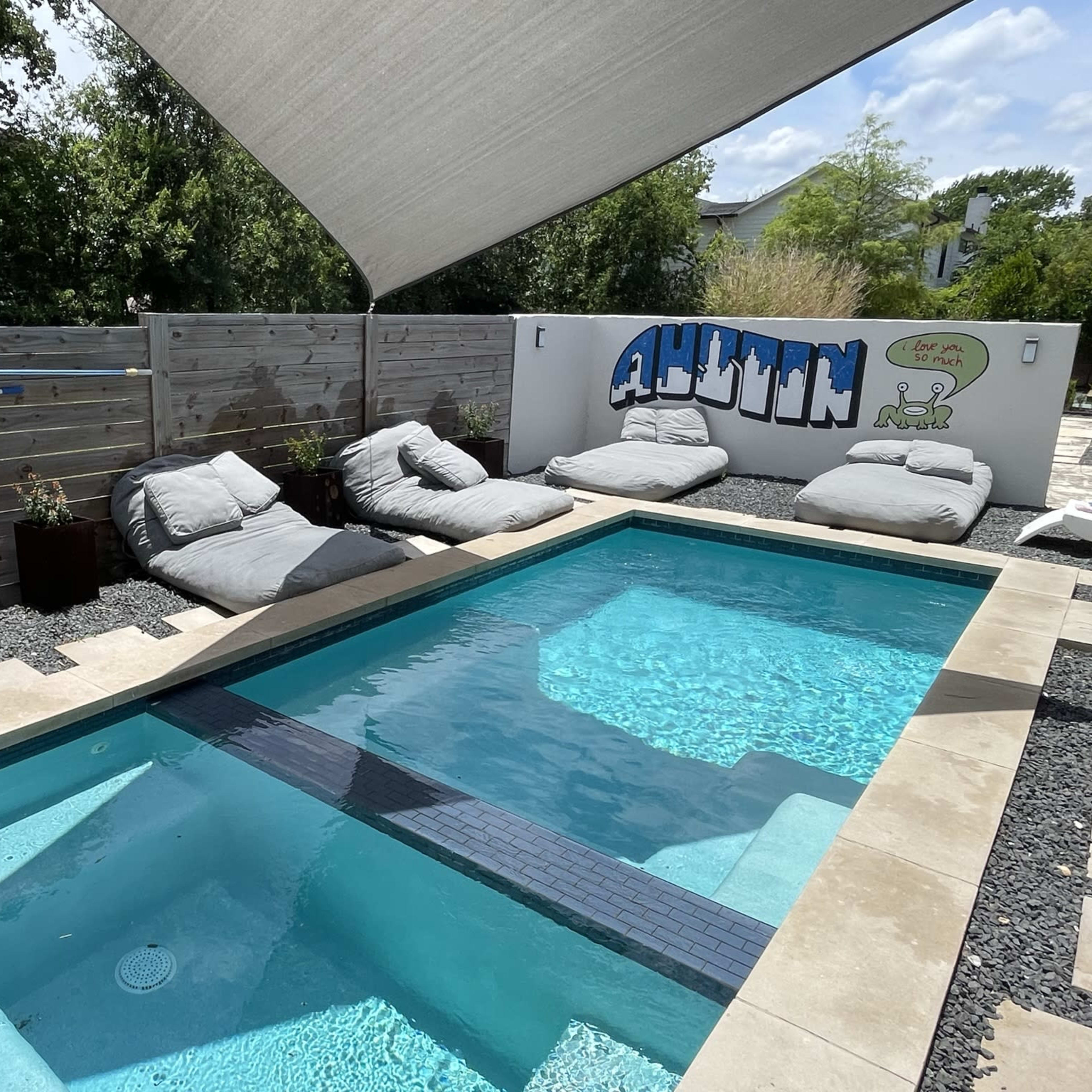 A rectangular swimming pool with a light stone deck is surrounded by large lounge cushions and a graffiti-style mural on a wooden fence.