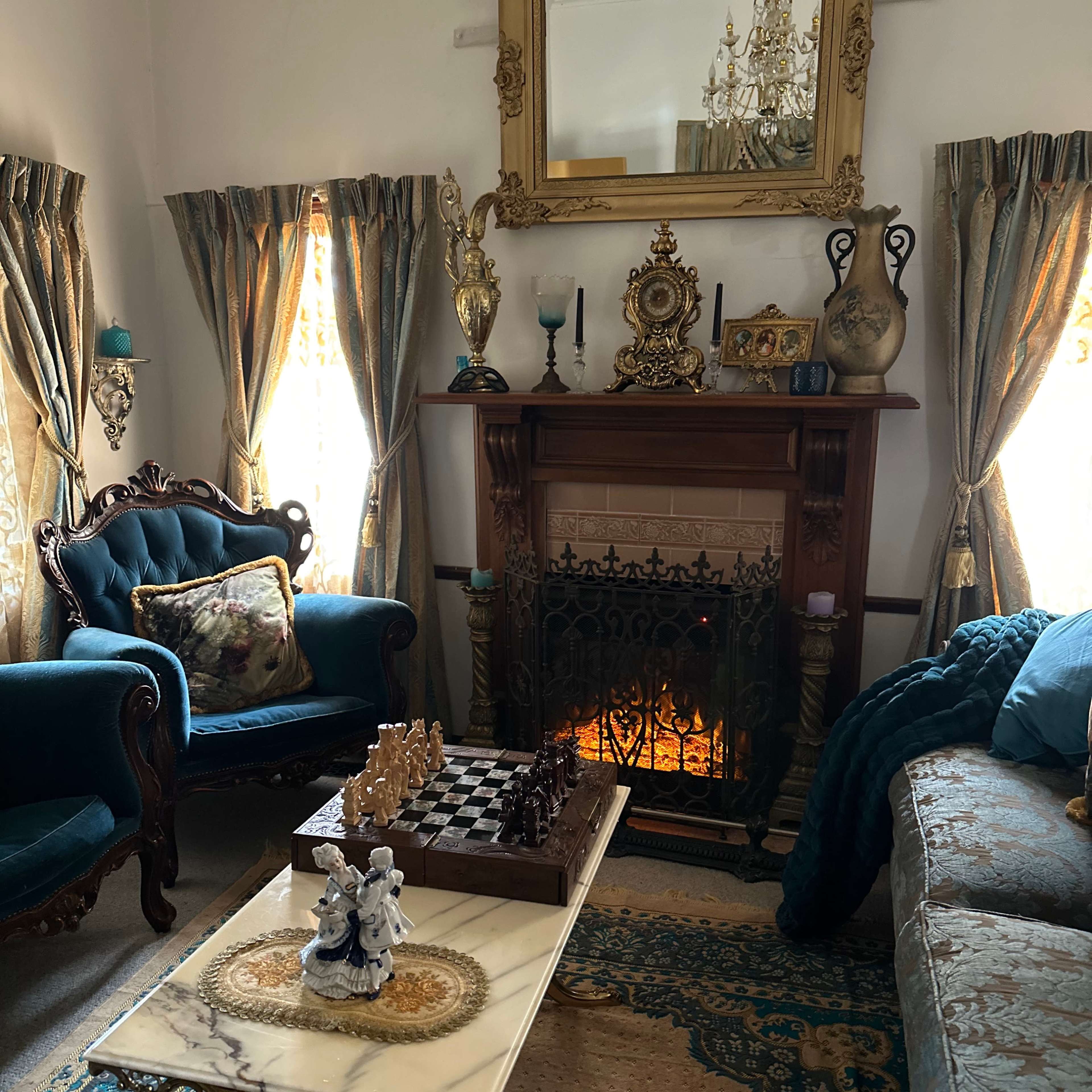 A cozy living room with a fireplace, elegant furniture, a chess board on a table, and ornate curtains framing the windows.