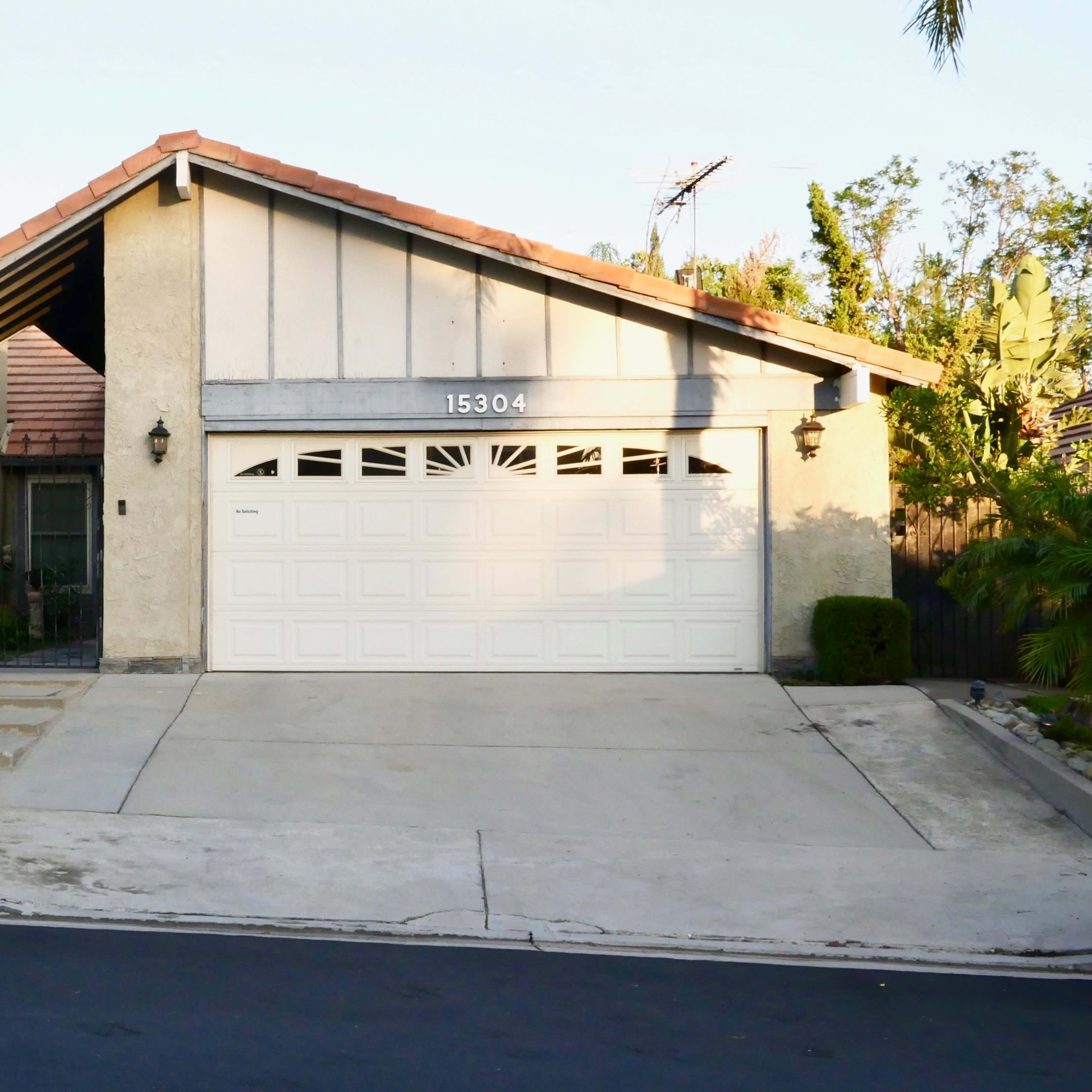 A single-story house with a two-car garage sits on a well-kept lawn surrounded by trees and shrubs.