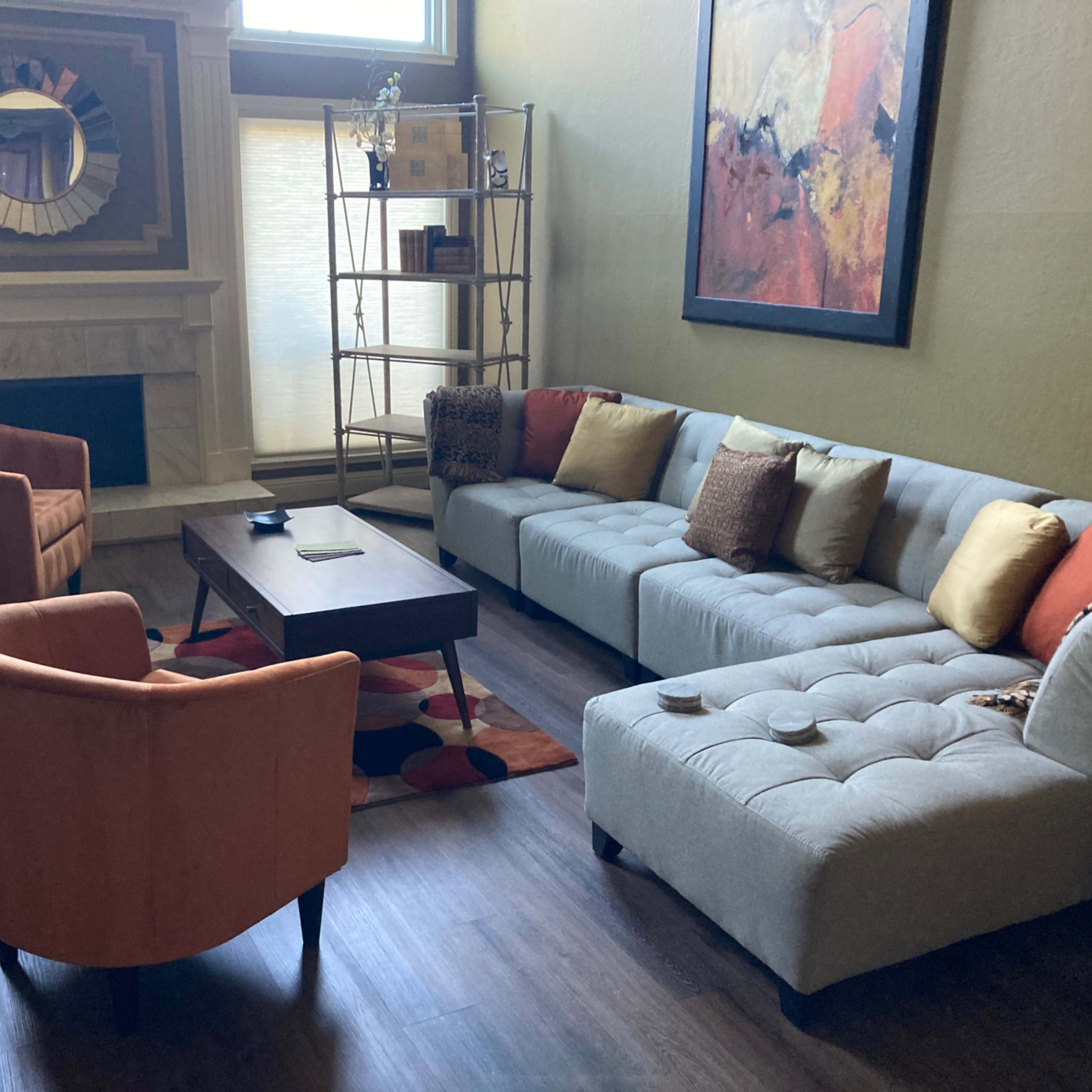 A living room with a gray sectional sofa, two orange armchairs, a wooden coffee table, and a bookshelf against a wall, all arranged around a fireplace.