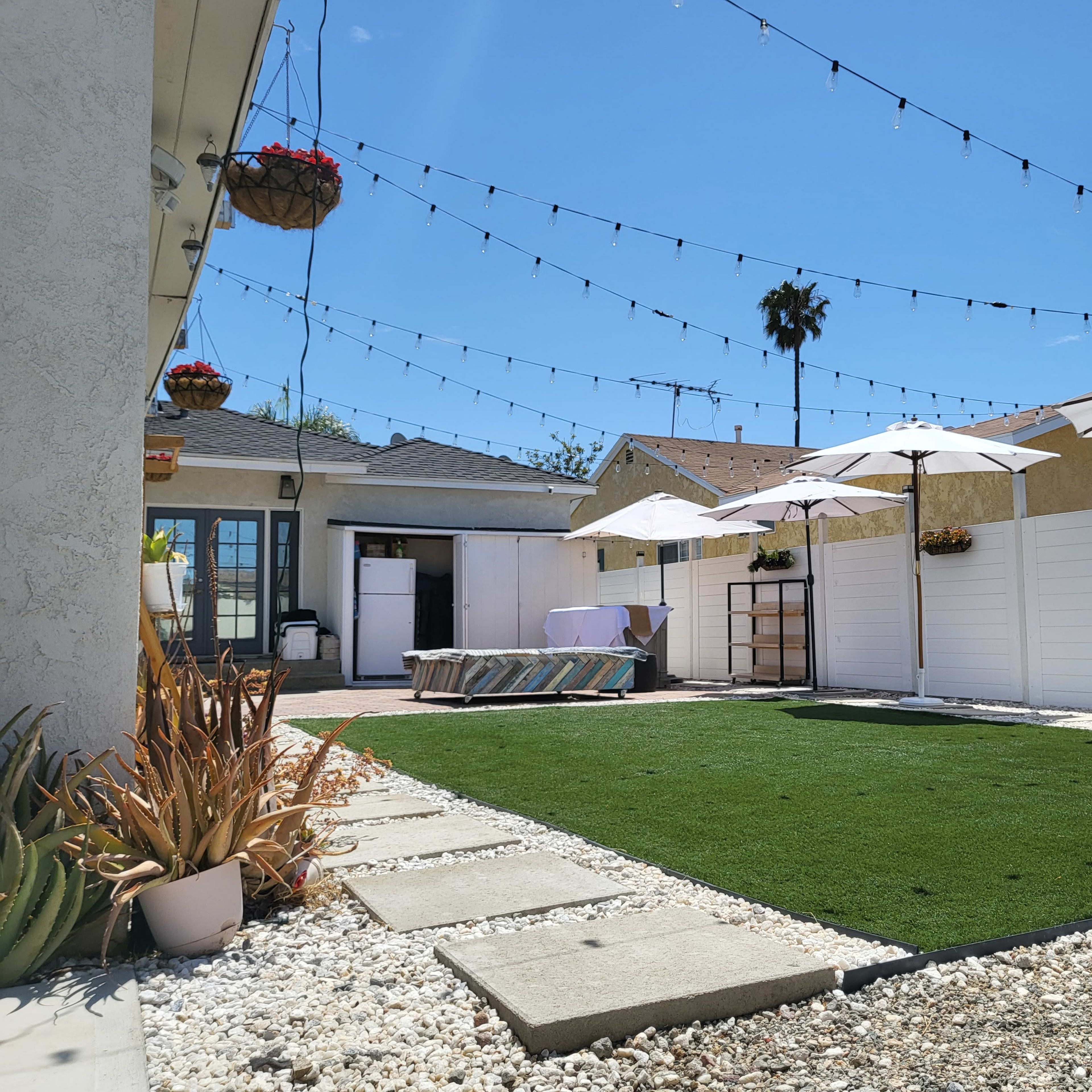 A landscaped backyard features artificial grass, decorative plants, and patio umbrellas, with string lights overhead and a clear blue sky.
