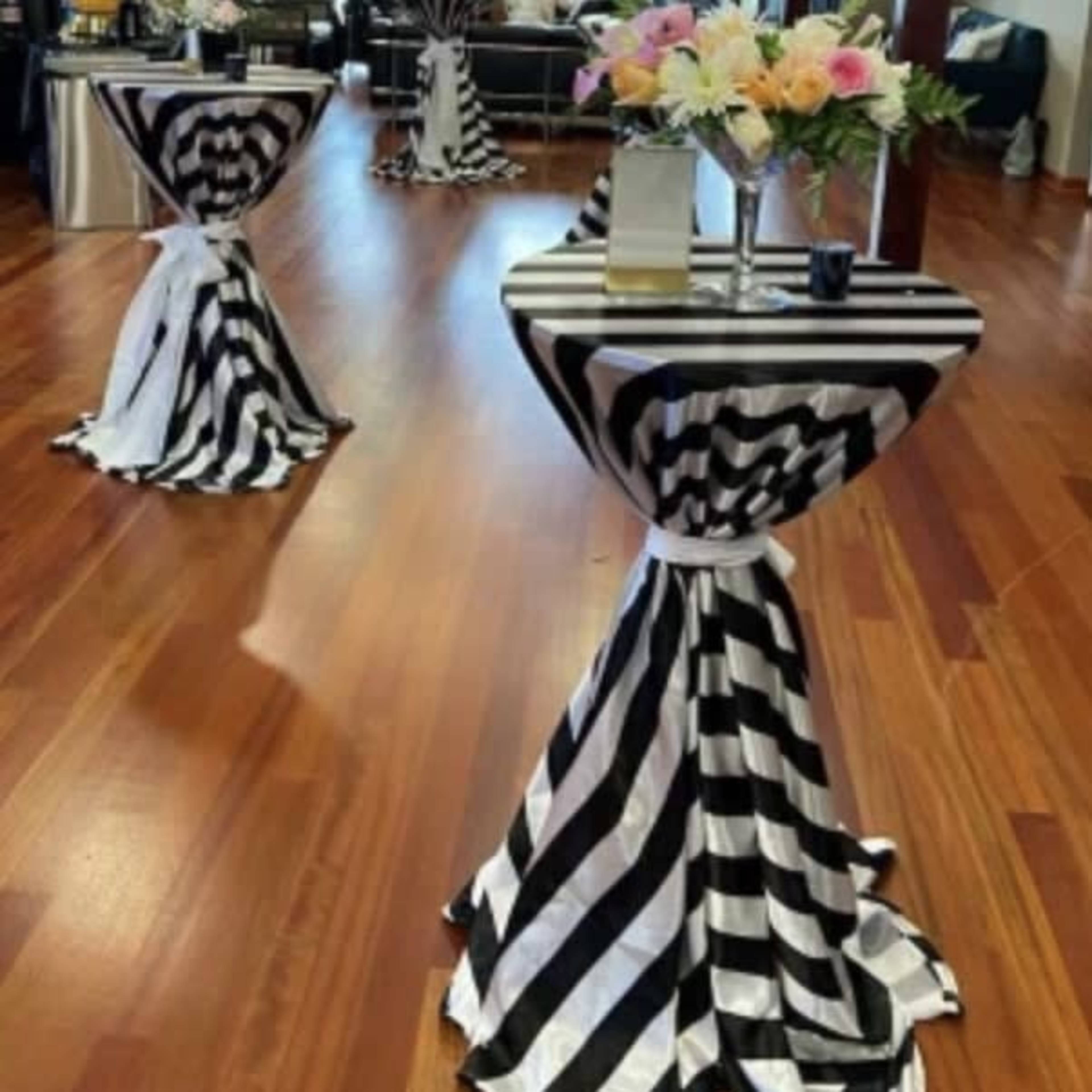 An event space with a polished wooden floor, decorated with striped black and white table covers and modern furniture against a backdrop of exposed brick walls and large windows.