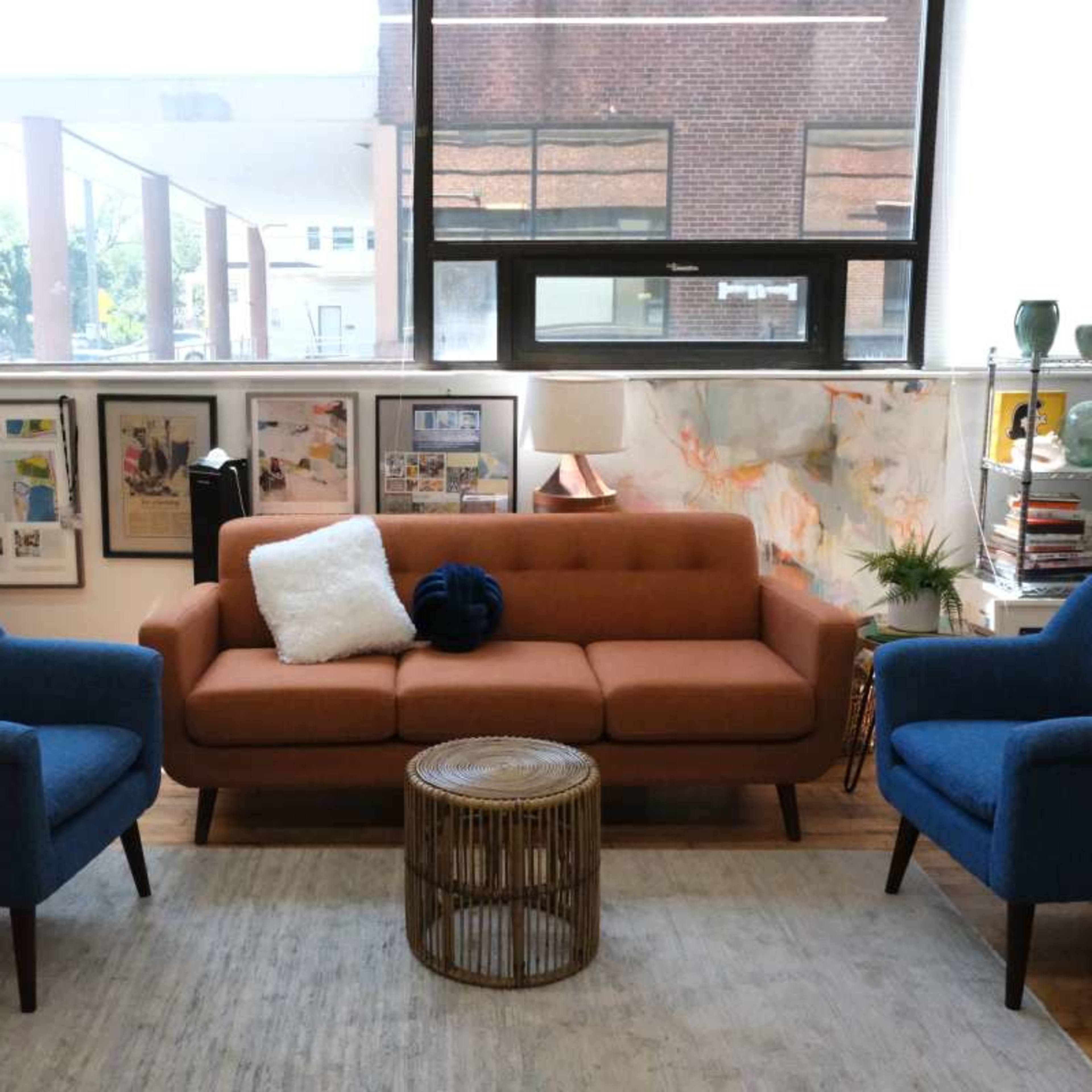 A modern living space featuring a brown sofa and two blue chairs arranged around a round, woven coffee table on a light rug.