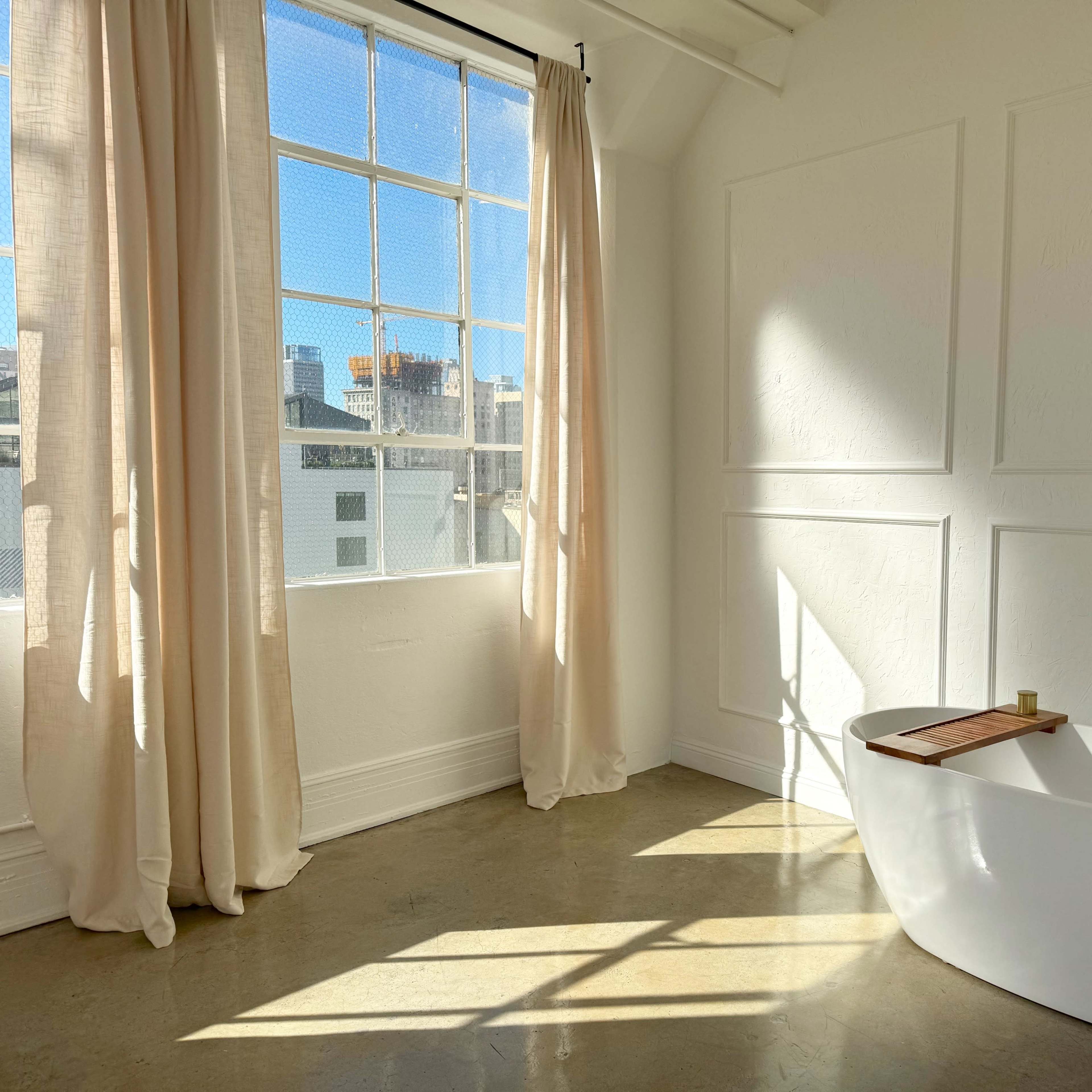 A bright bathroom with a freestanding white bathtub near a large window adorned with light curtains.