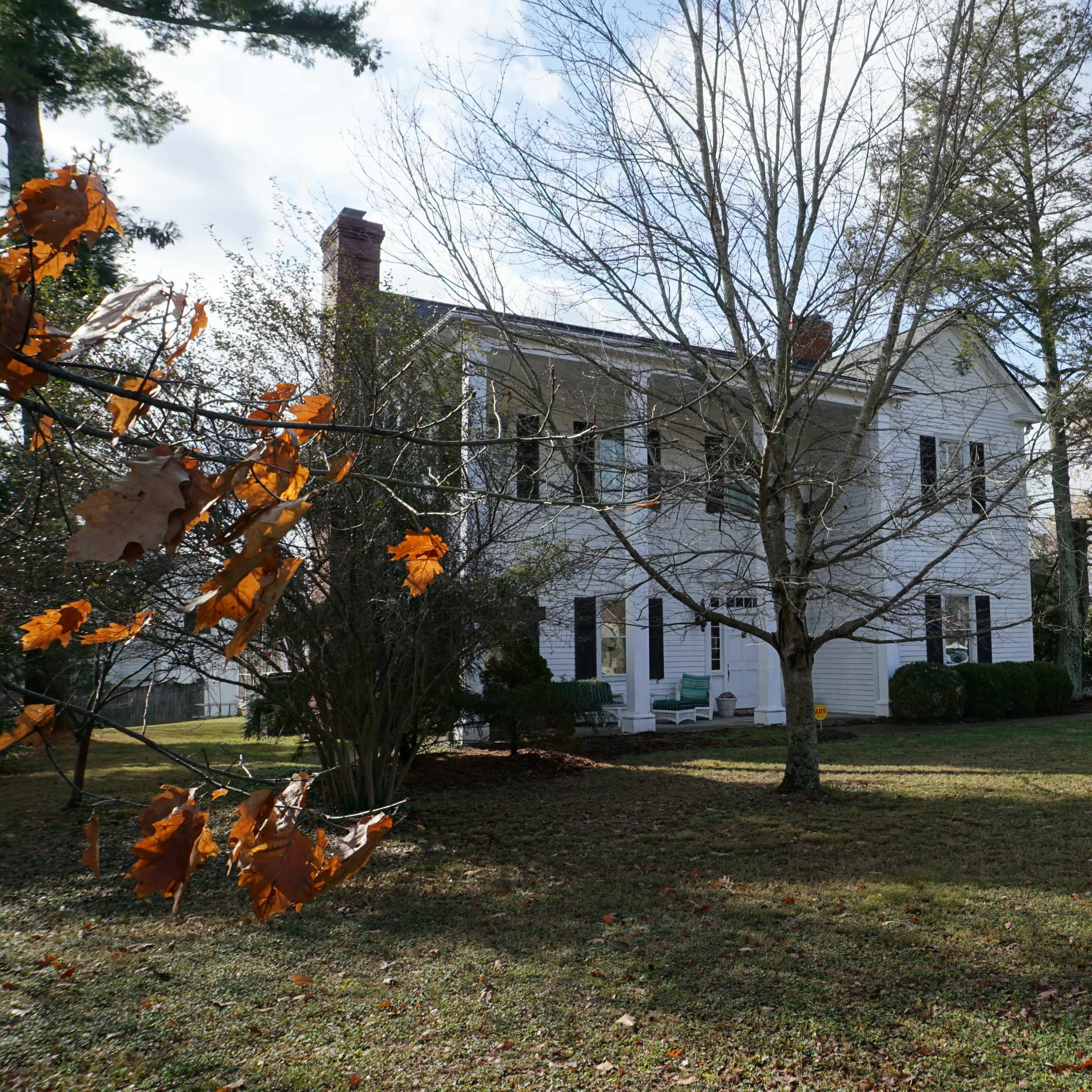 A two-story white house stands among trees on a grassy lawn, with autumn leaves scattered on the ground.