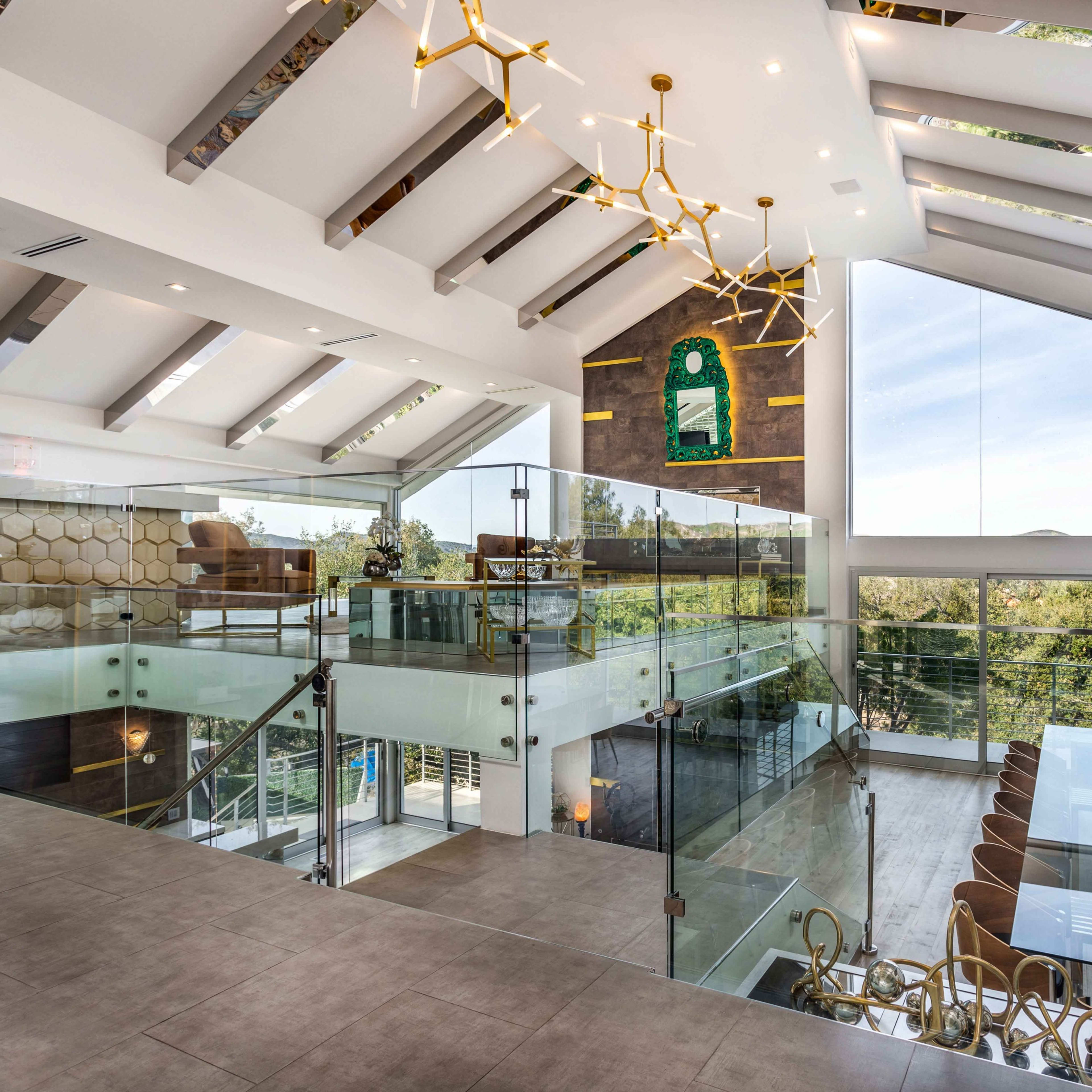 A spacious, modern interior features a glass dining table surrounded by elegant chairs, with a view of greenery through large windows and a staircase with a glass railing.