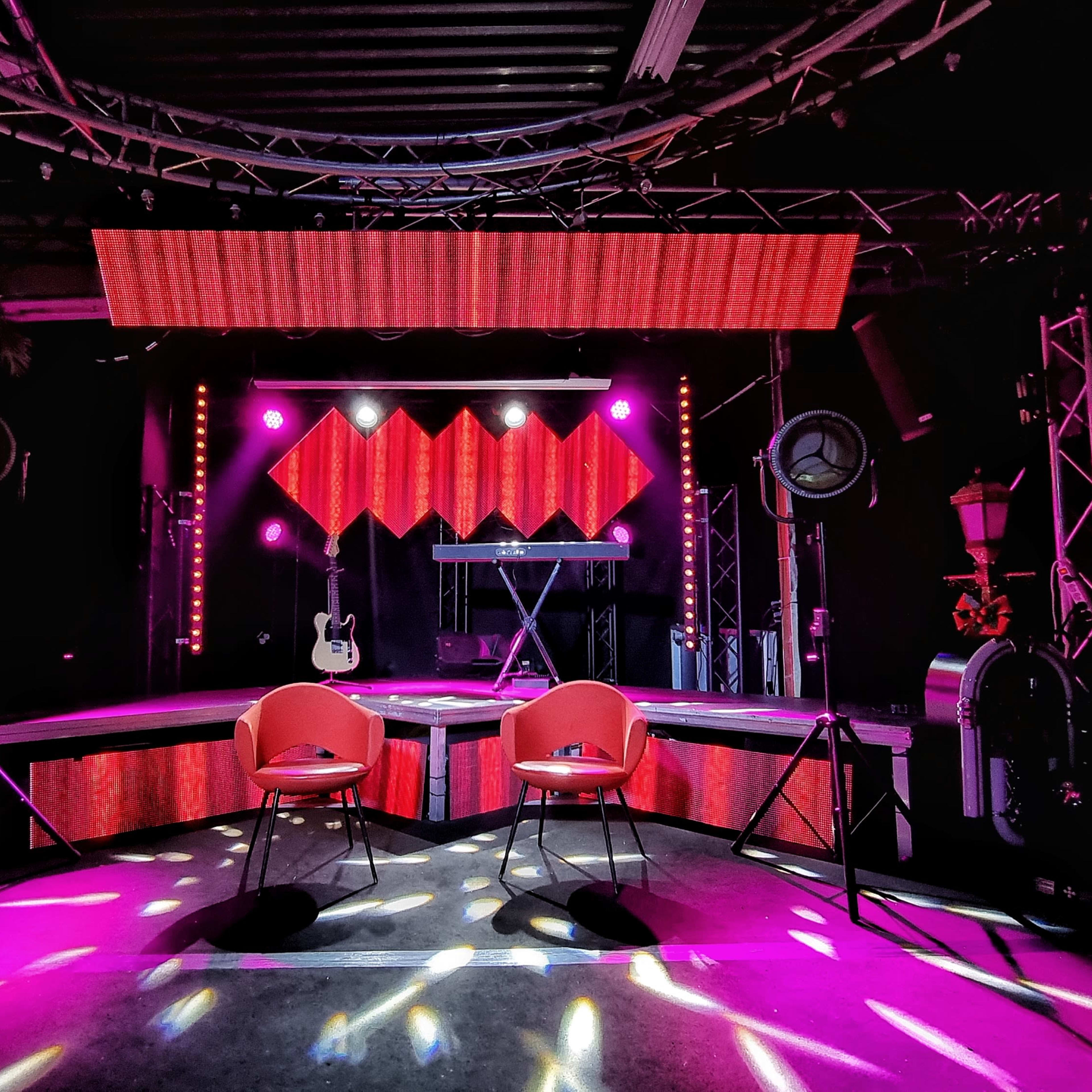 A stage setup featuring two chairs, a guitar, and a keyboard under vibrant stage lights and a decorative backdrop.