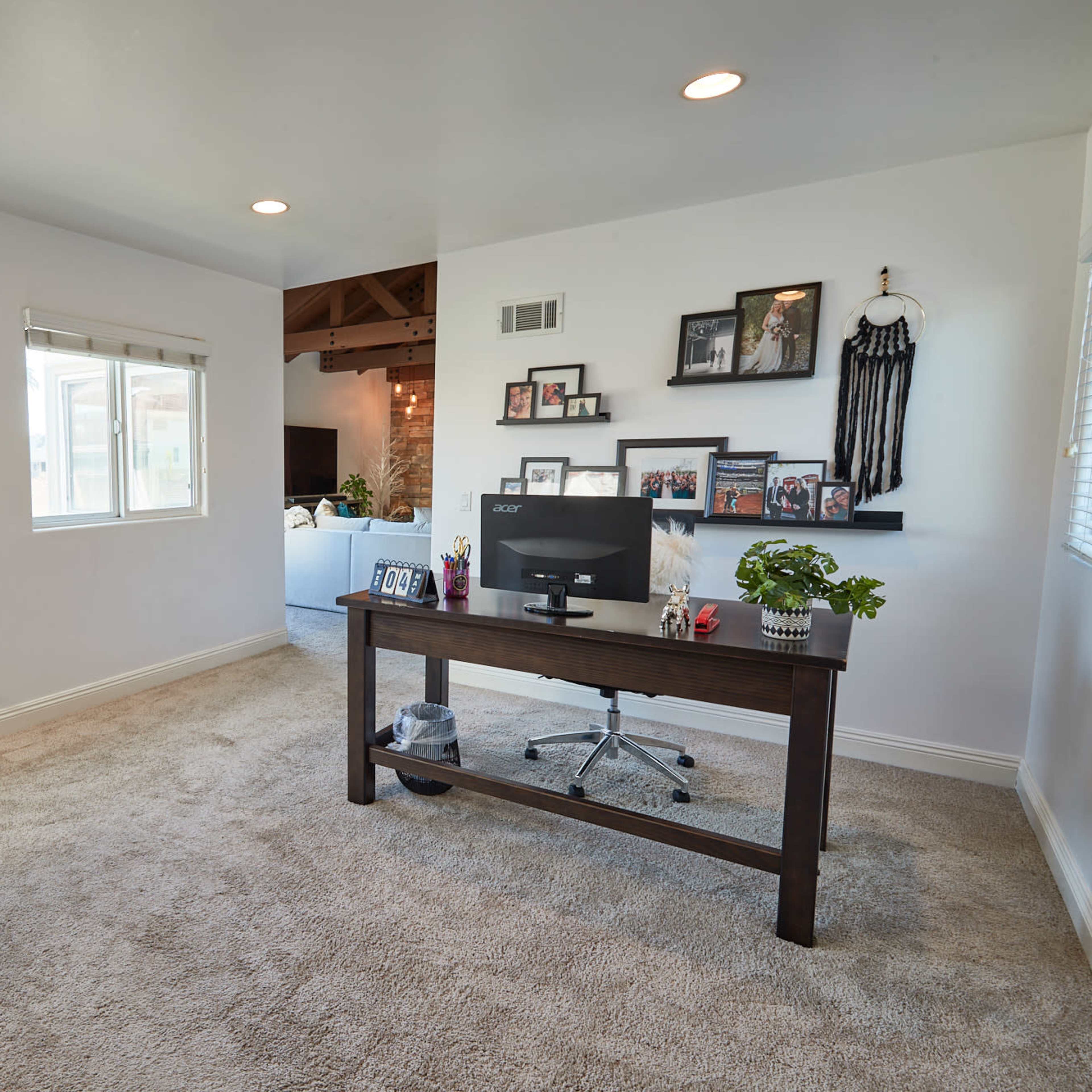 A home office features a wooden desk with a computer, surrounded by windows and decorated with family photos.