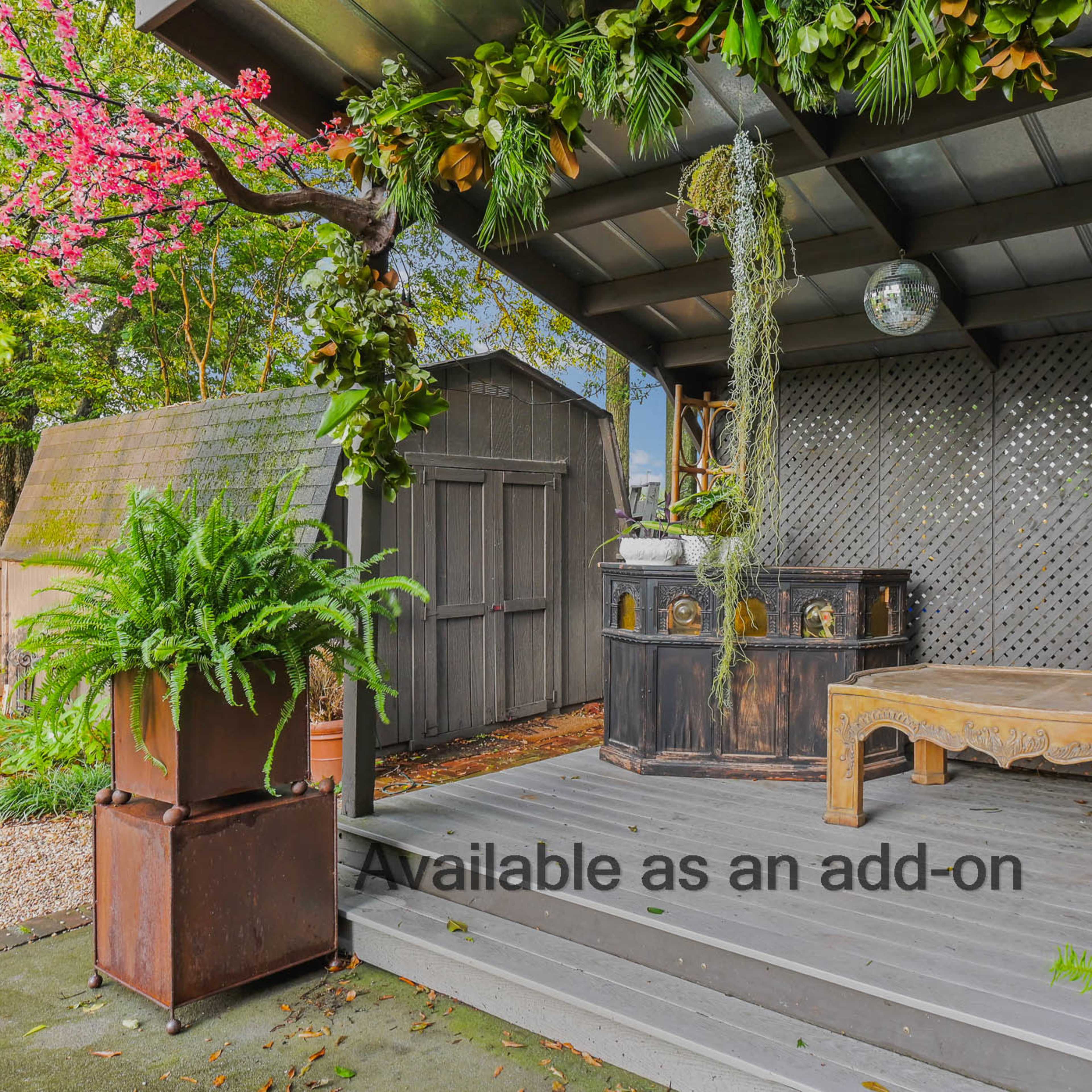 A cozy outdoor space with a wooden bar, a table, hanging greenery, and a small shed in the background.