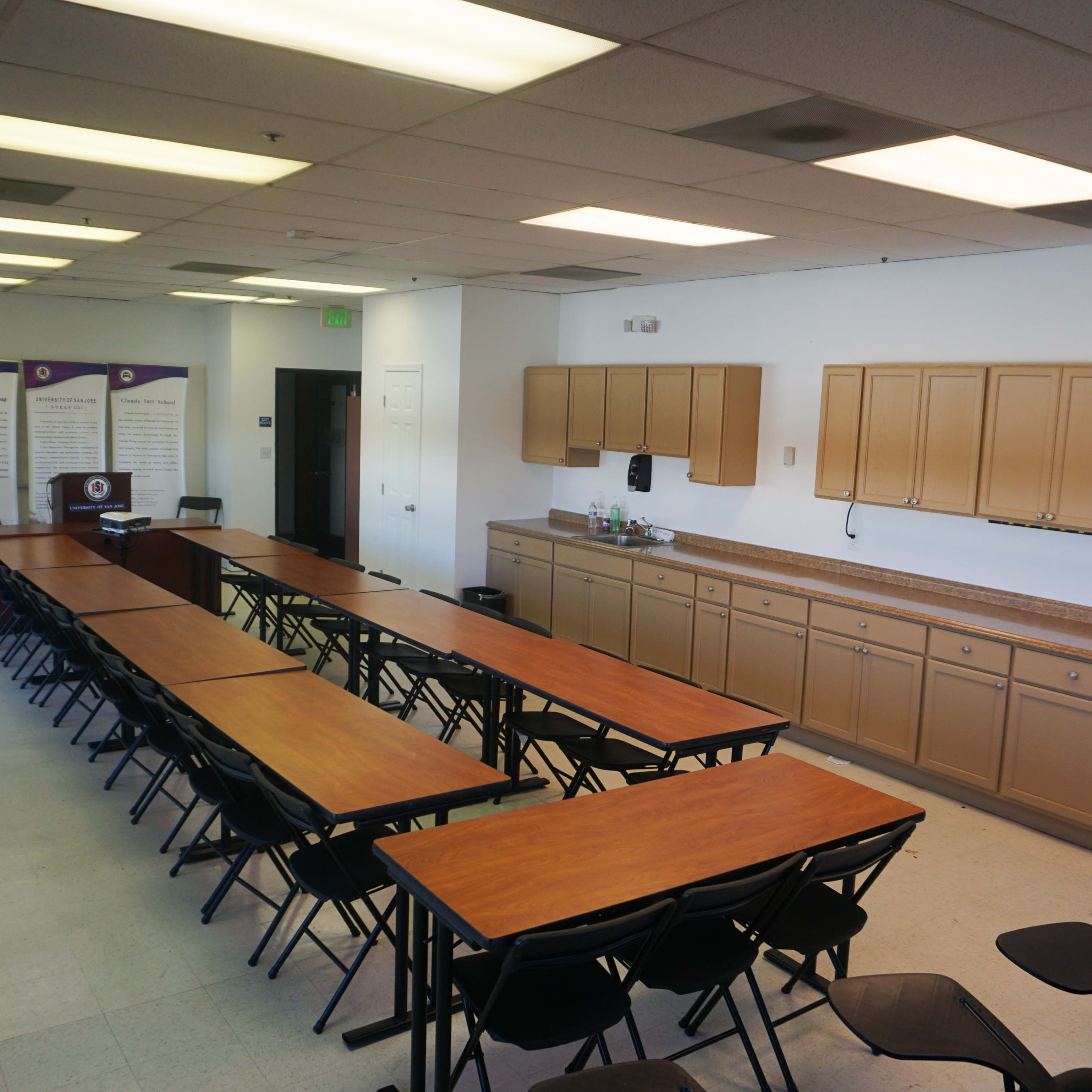 A spacious meeting room with several rows of rectangular tables and chairs arranged to facilitate presentations, along with a kitchen area featuring cabinets and a countertop.