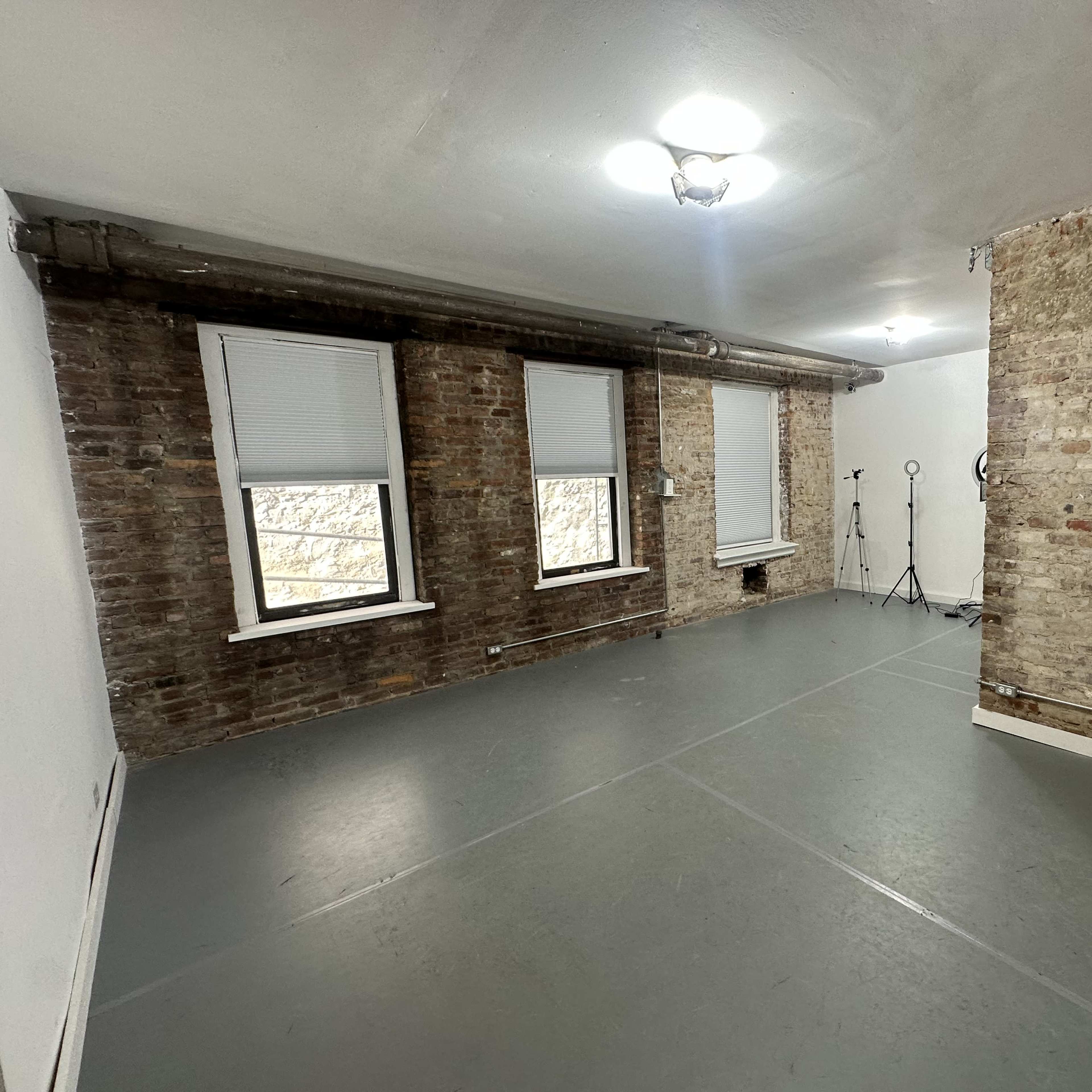 A spacious room with exposed brick walls, large windows, and a gray floor, featuring minimal furniture and a lighting stand in one corner.