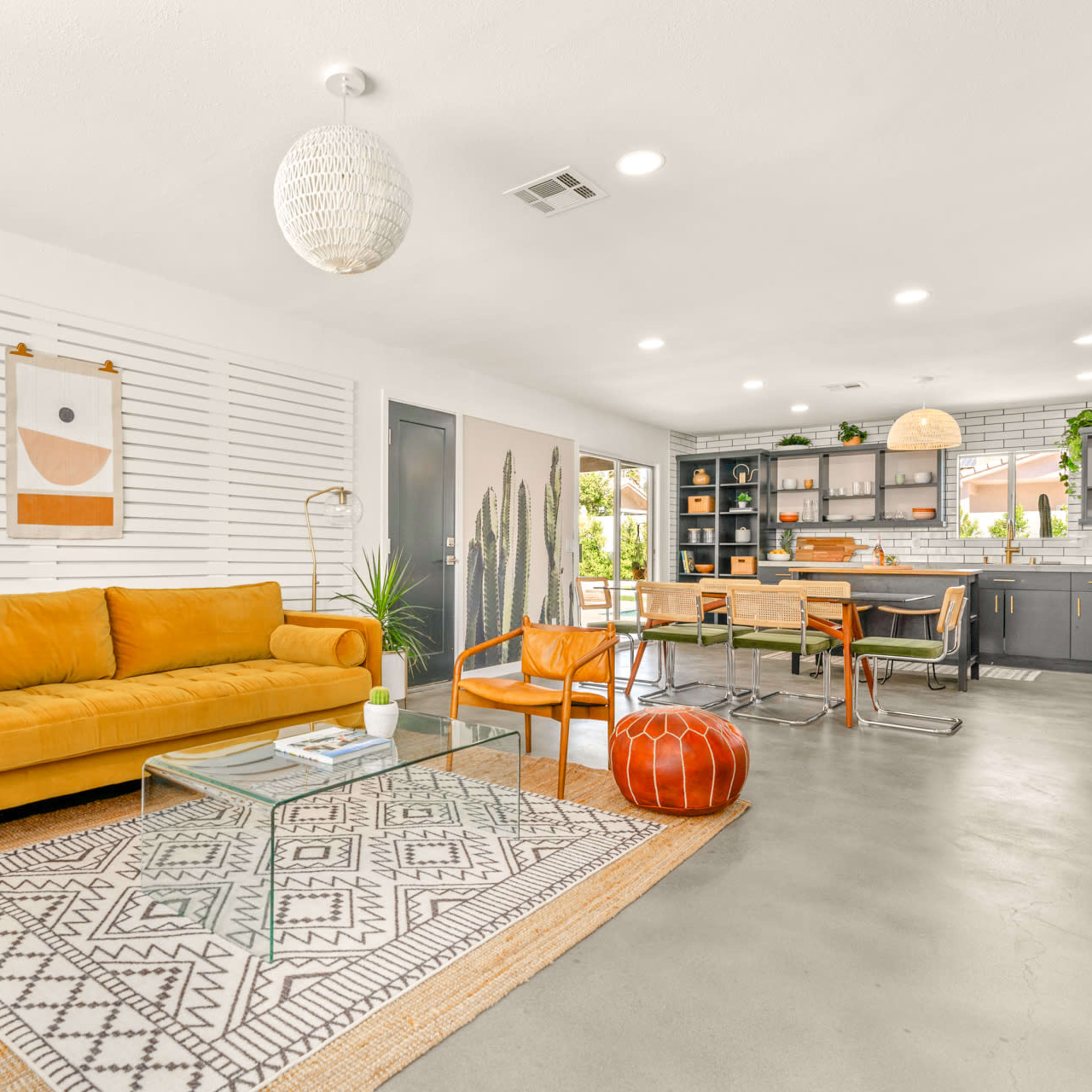 A modern living room with a mustard-colored sofa, a glass coffee table, and an open dining area featuring a black kitchen and wooden dining chairs.