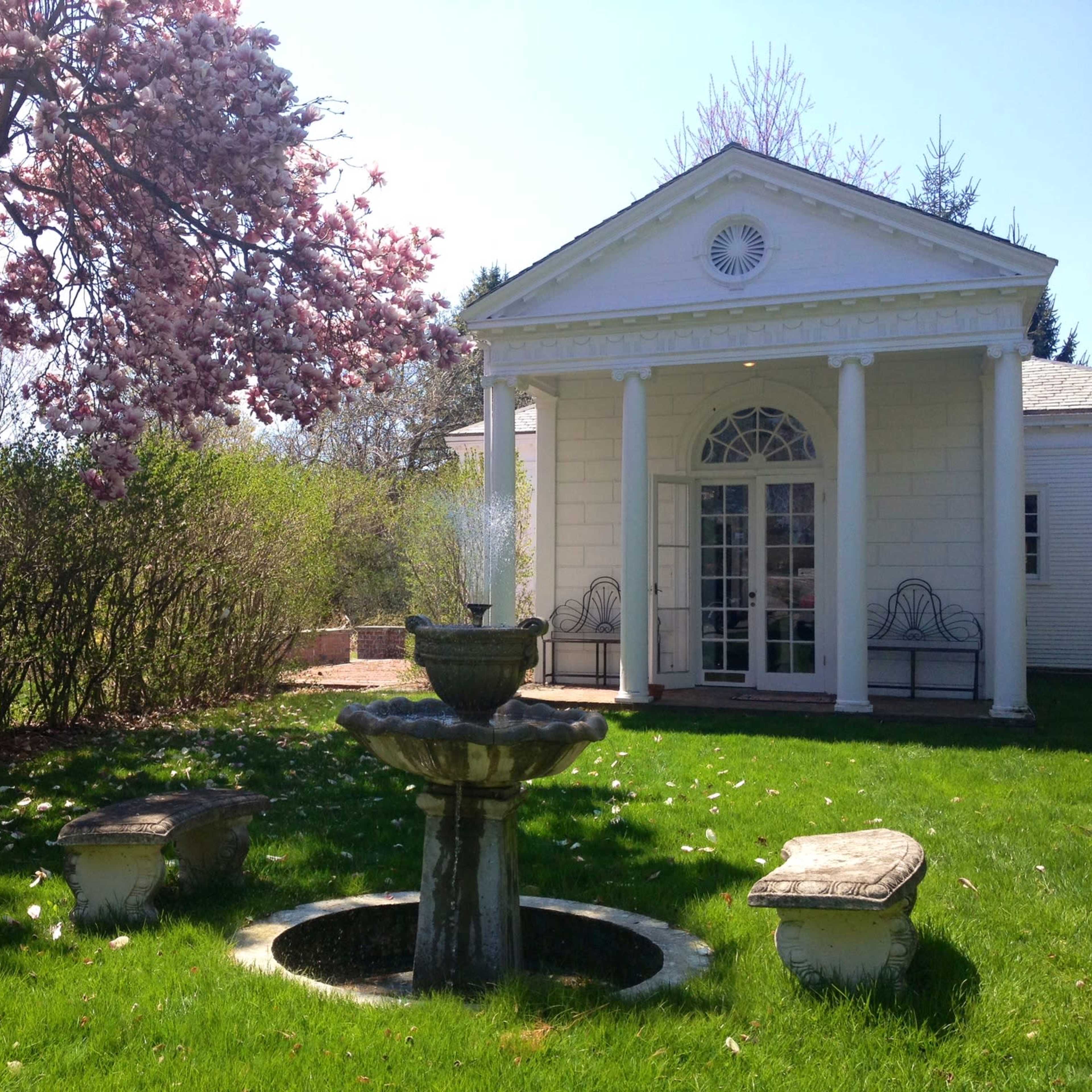 A fountain sits in a grassy area surrounded by flowering trees and a white building with columns.
