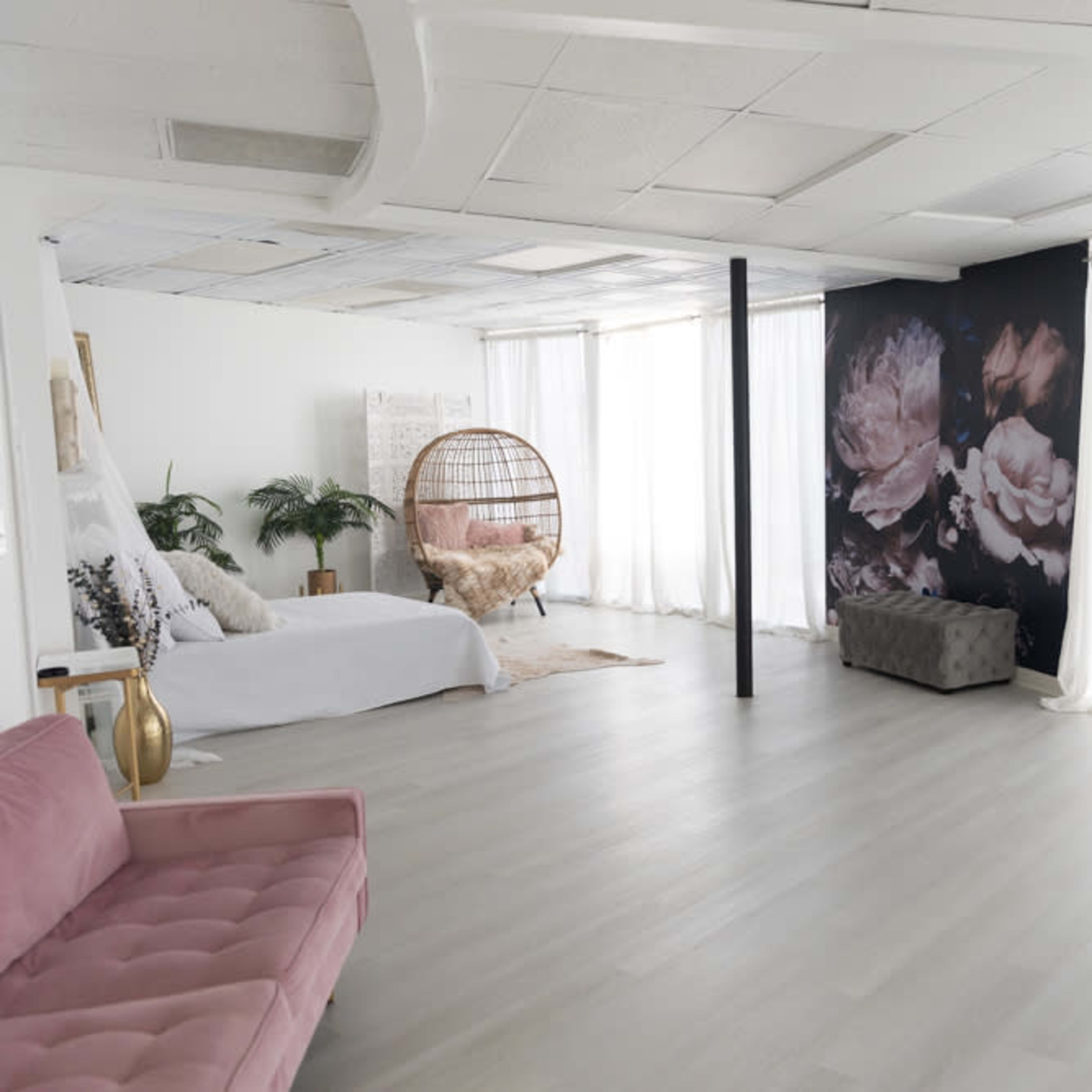 A spacious, well-lit room featuring a white sofa, a decorative hanging chair, a floral mural, and large sheer curtains.