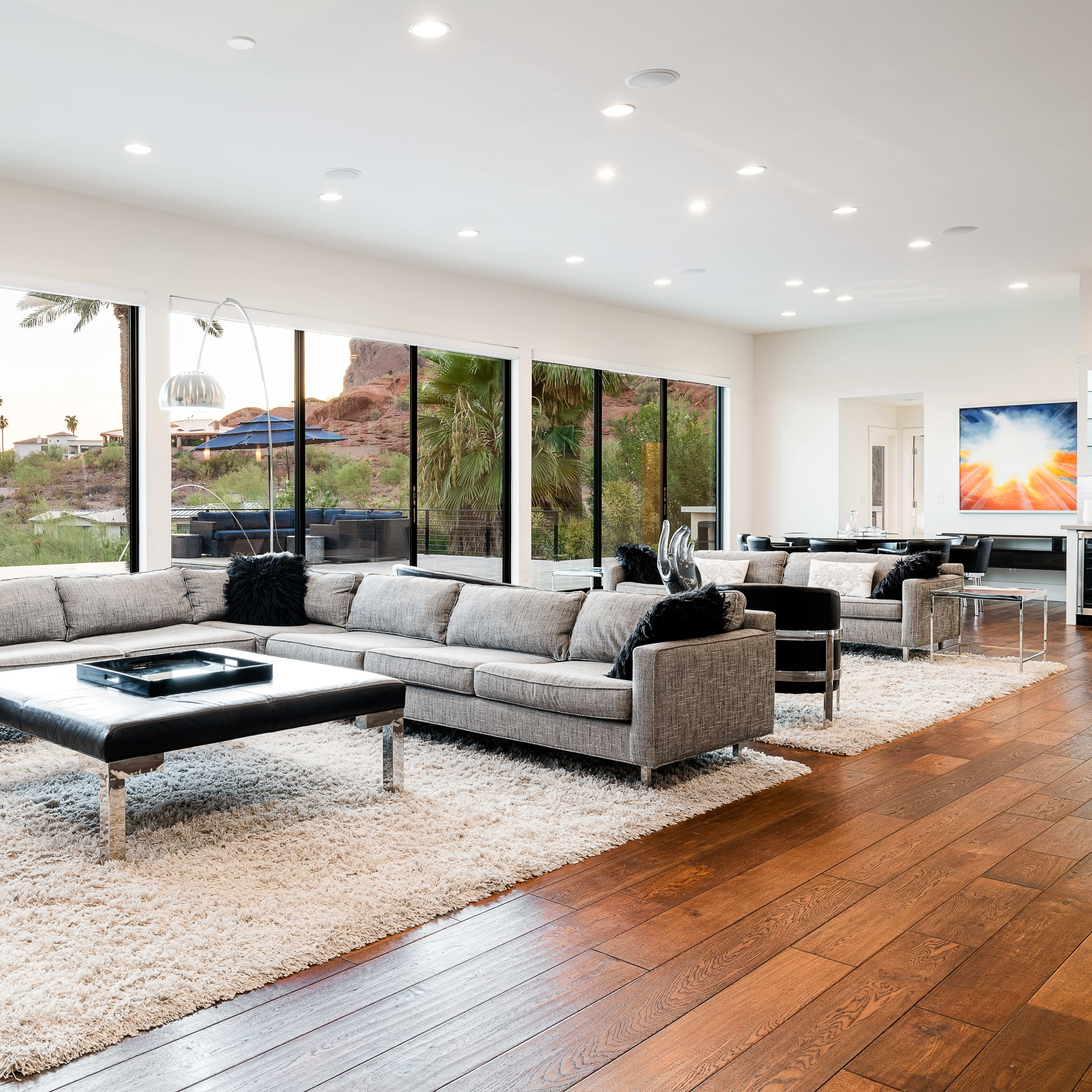 The living room features a large gray sectional sofa, a black coffee table, and expansive windows that provide views of palm trees and a natural landscape.