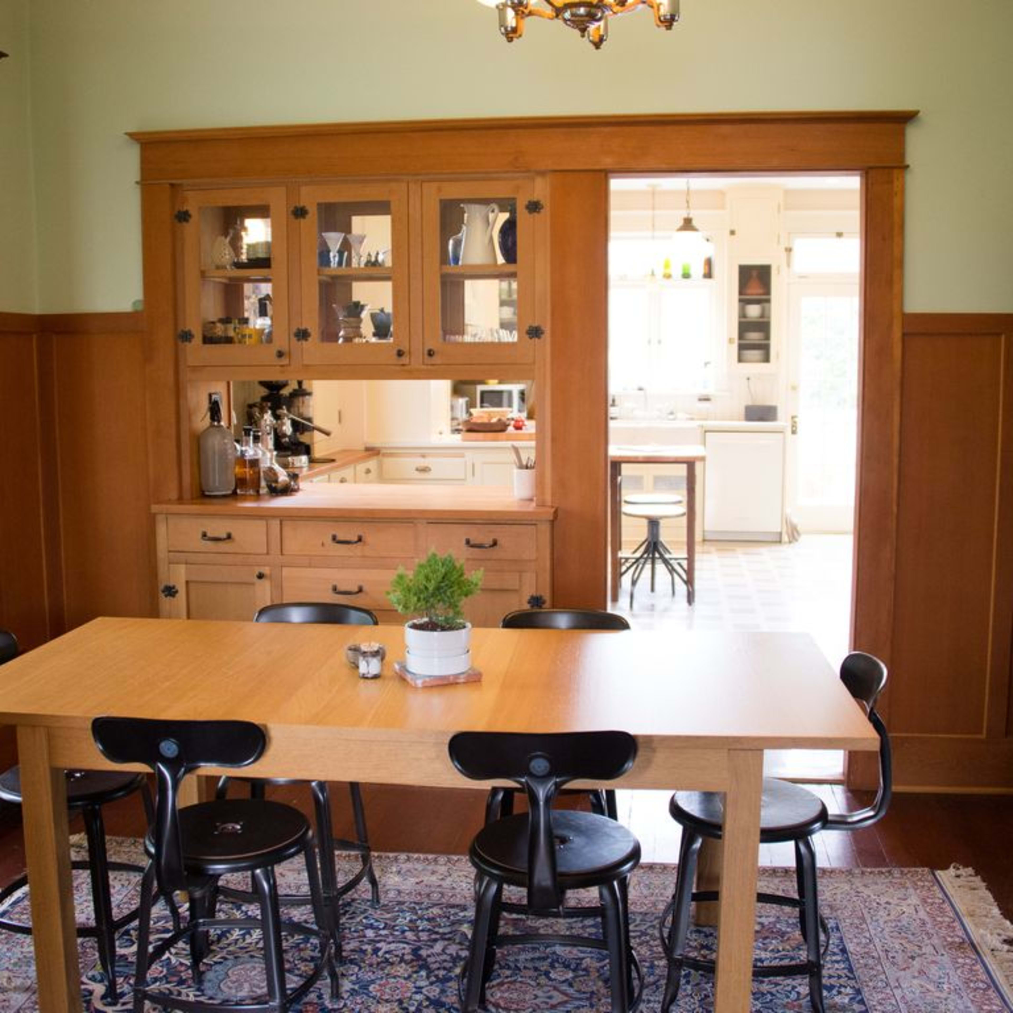 A wooden dining table with black chairs is set in a room featuring light green walls and wooden cabinetry, with a doorway leading into a kitchen.
