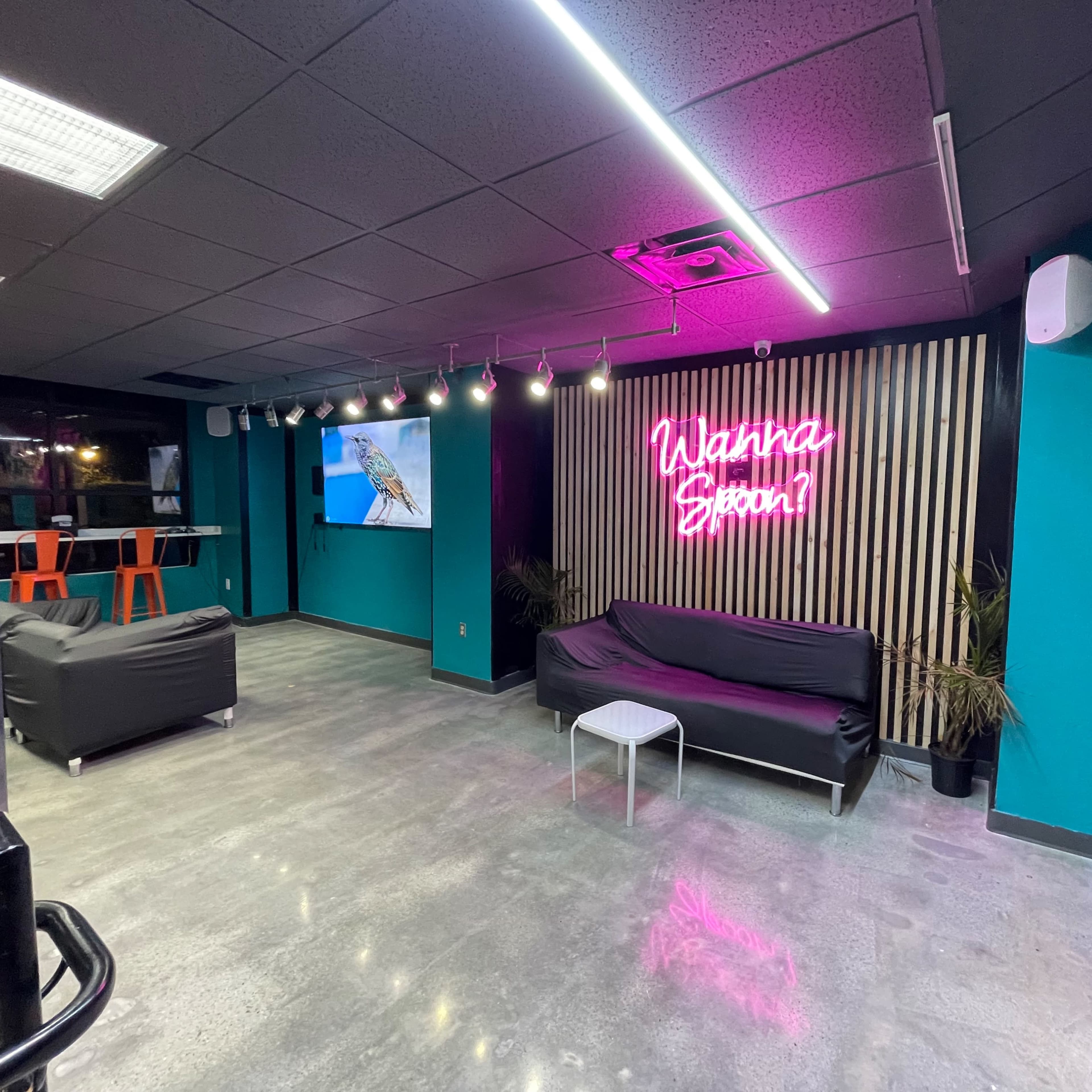 A modern lounge area with black sofas, a white table, and a neon sign on the wall saying "Wanna Blook?" illuminated in pink light.