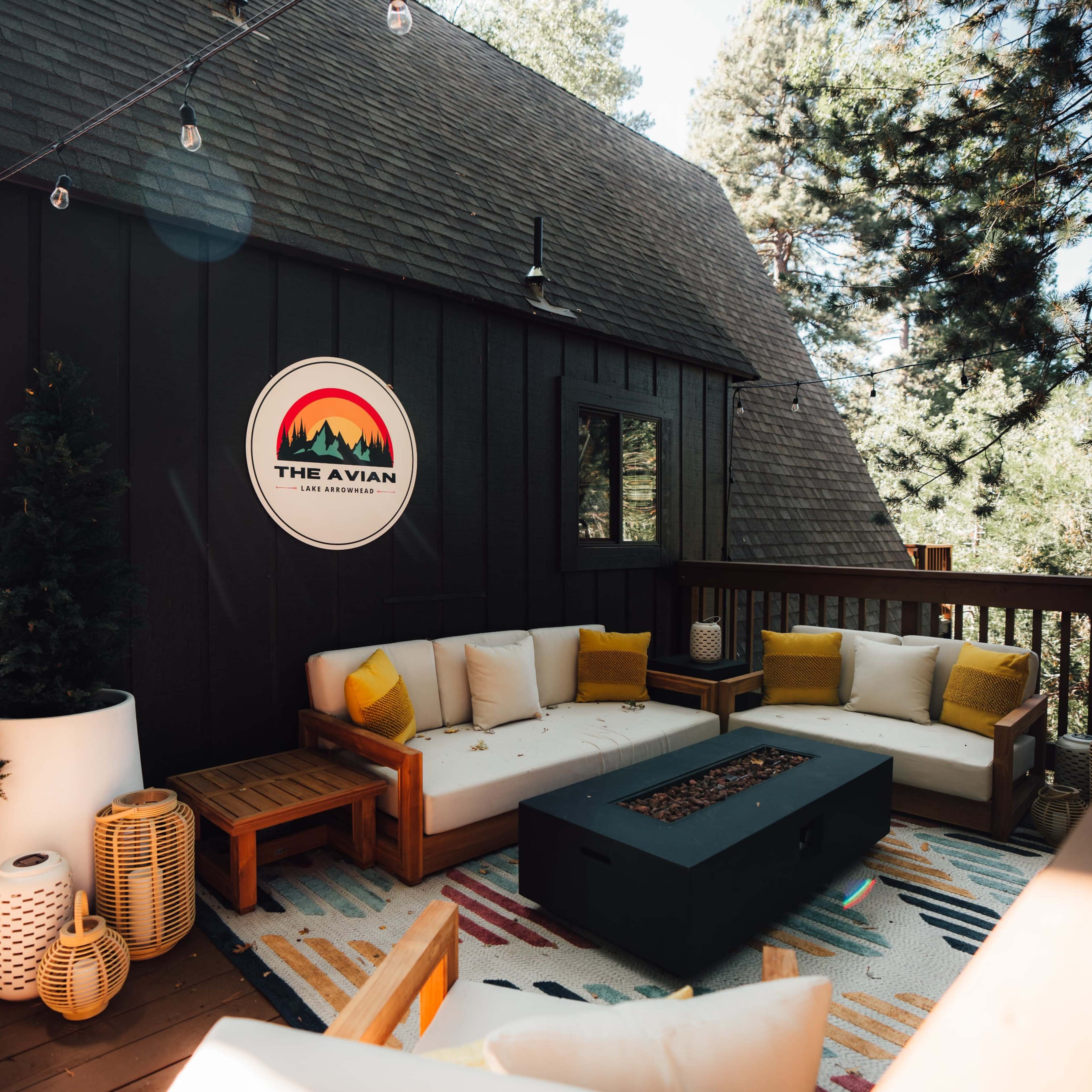 A cozy outdoor seating area features a sectional couch, a coffee table with a fire pit, and decorative pillows, set against a backdrop of trees and a dark wood-paneled cabin.