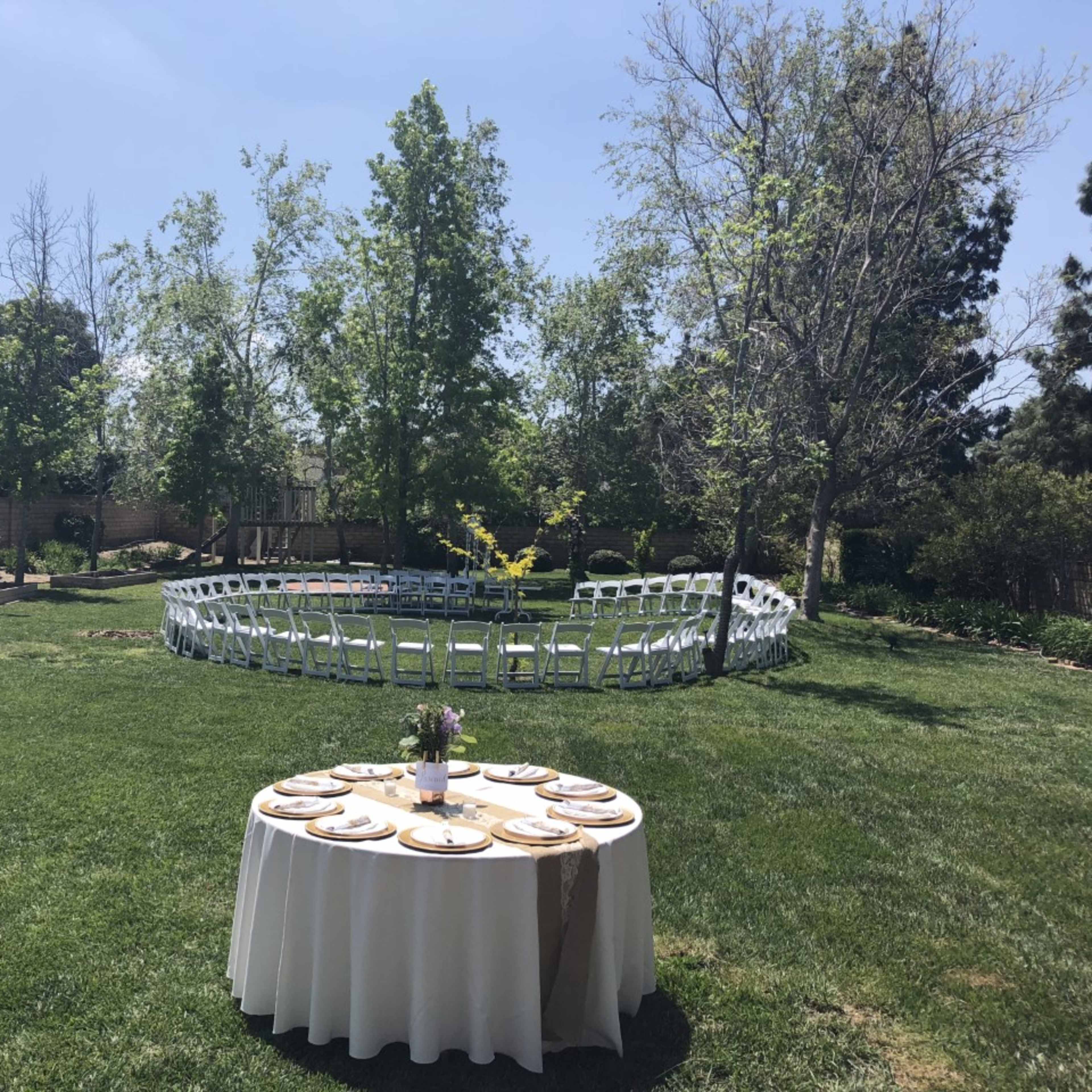 A circular arrangement of white chairs is set up on a green lawn, with a table adorned with plates and a floral centerpiece in the foreground.